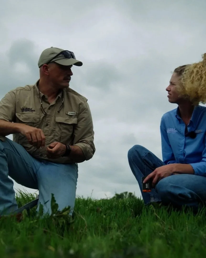 Two people, a man and a woman, are outdoors on grass, engaging in a conversation. The man is wearing a beige shirt, a cap, and sunglasses on his head. The woman has curly blonde hair and is wearing a blue shirt and jeans.