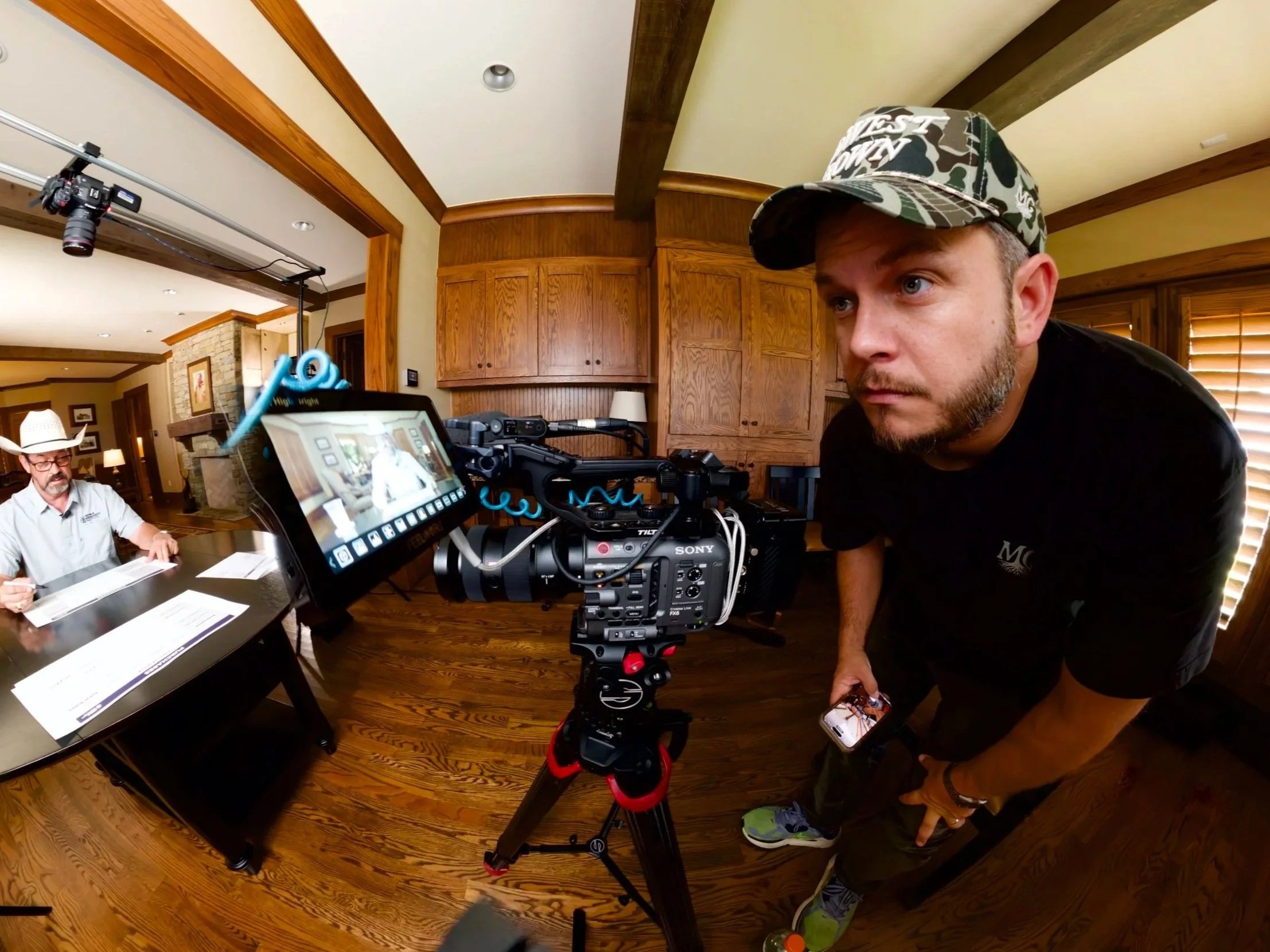 A man in a camouflage cap looking into a professional camera camera mounted on a tripod, with another man wearing a cowboy hat in the background in a cozy wooden-paneled room.