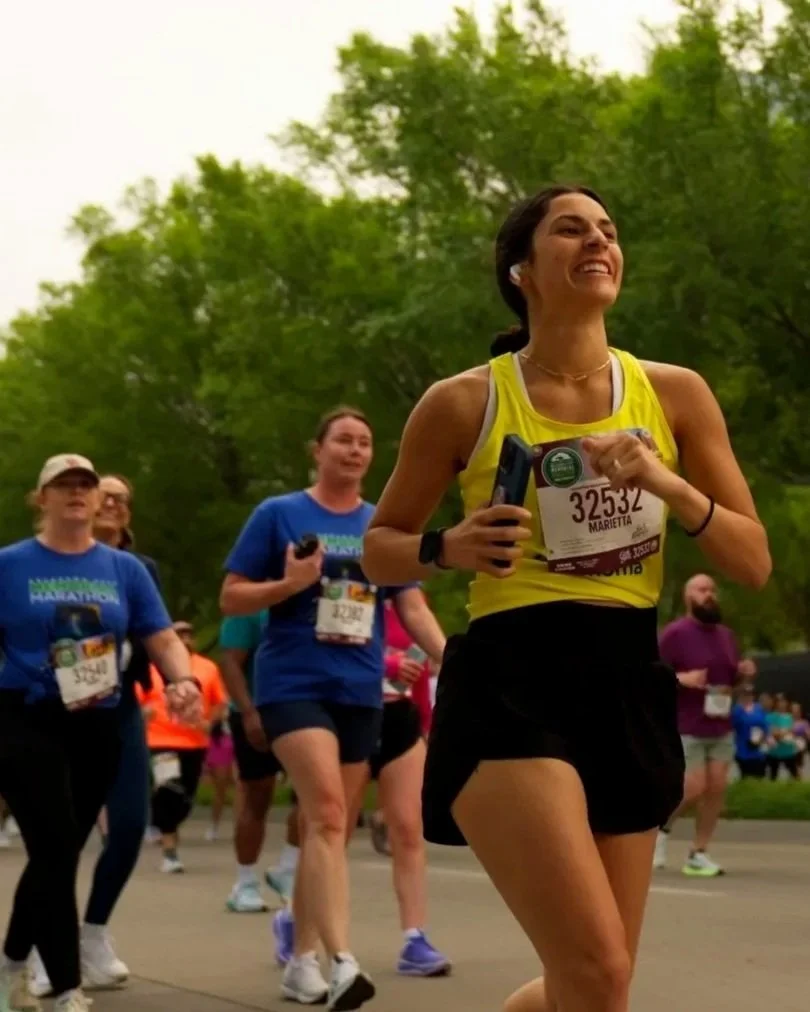A group of people participating in the Oklahoma city marathon, running outdoors in a park with green trees in the background. The woman in the foreground is smiling, wearing a yellow tank top, black shorts, and a race bib numbered 32532.