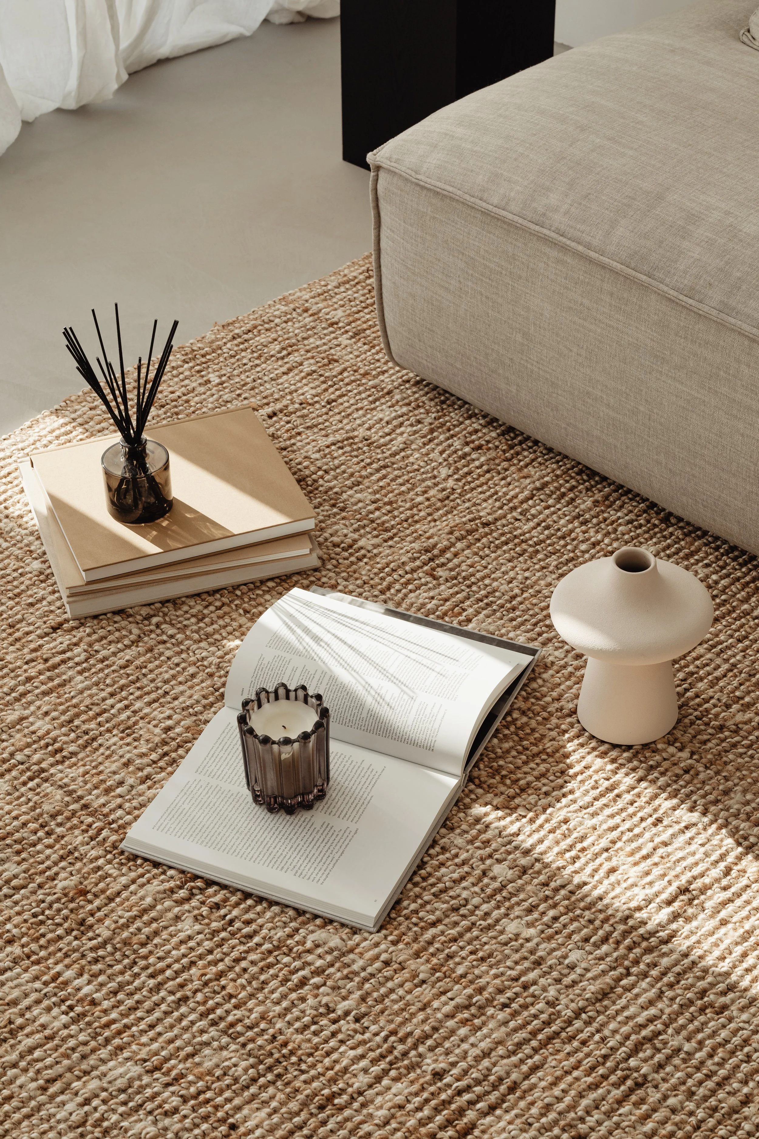 A beige textured area rug with a tray holding a candle and a stack of books, a white open book with text, a decorative glass candle holder, and a beige ceramic vase, next to a light-colored upholstered sofa in a cozy living space.