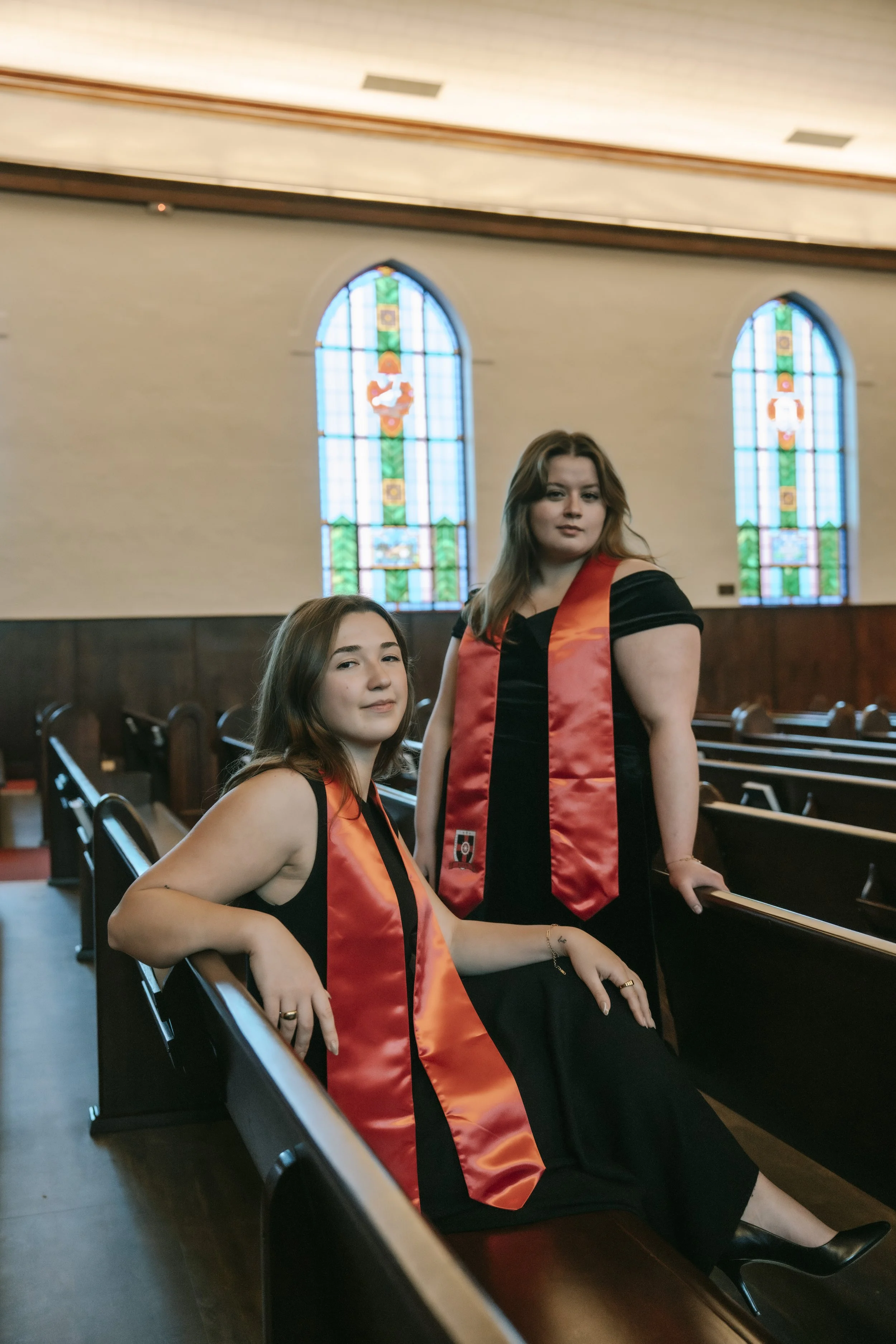 Two women in graduation gowns and stoles inside a church, with stained glass windows behind them.