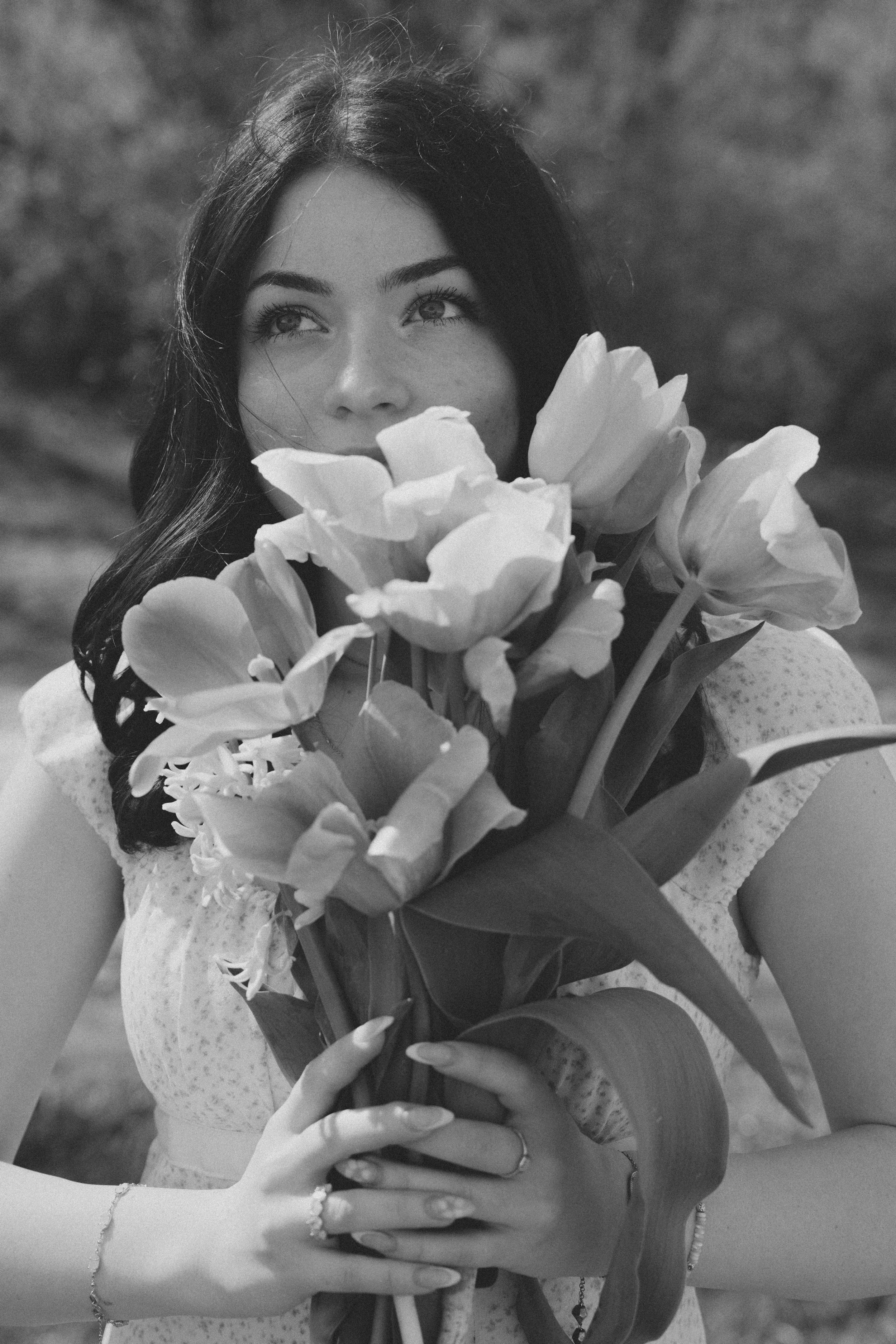 A woman with long dark hair holding a bouquet of large flowers, looking to the side, outdoors in black and white.