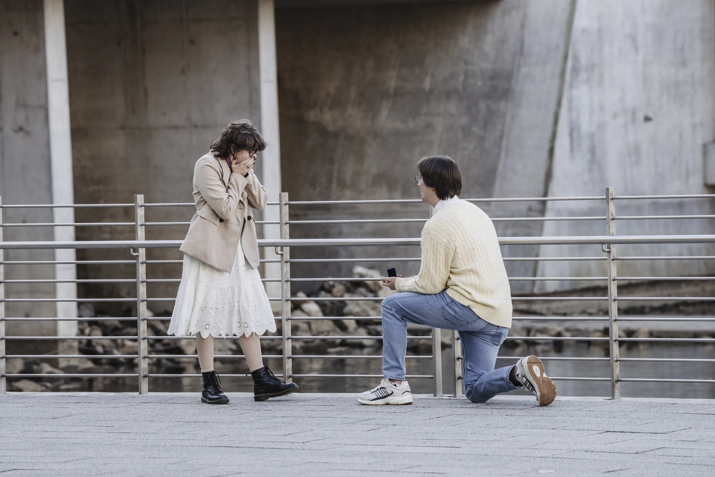 A man is kneeling on one knee, proposing to a woman on a bridge with a concrete background. The woman looks surprised and emotional, covering her face with her hands.