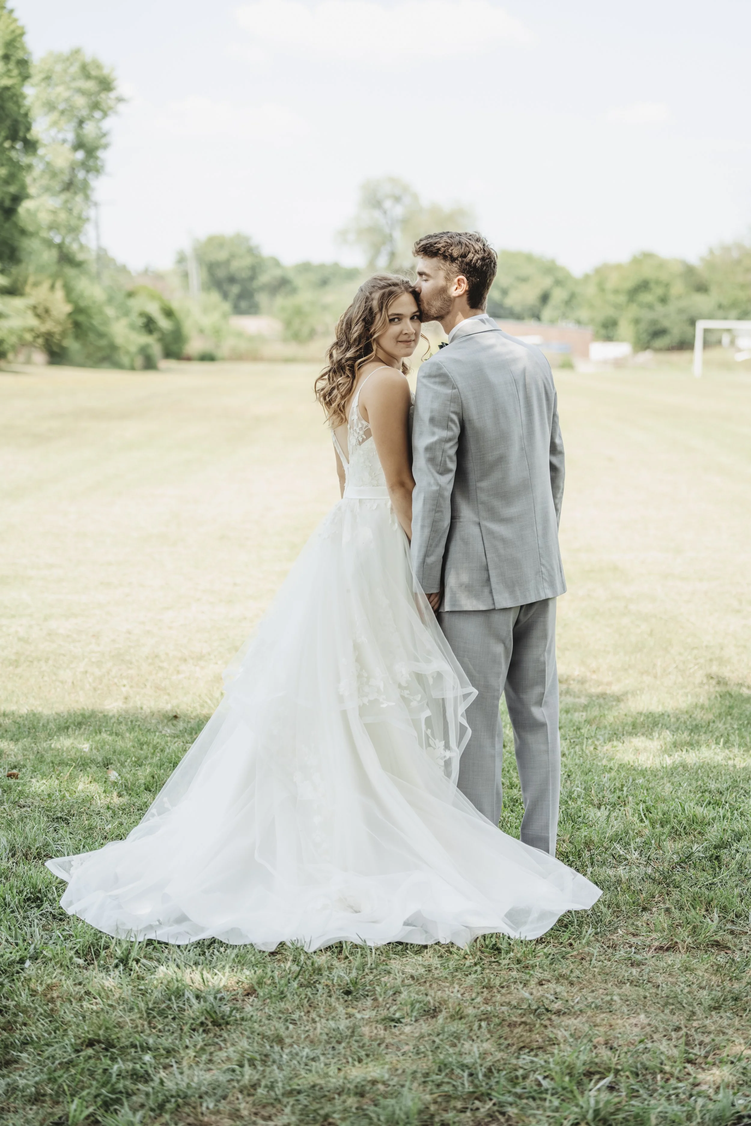 A bride and groom standing on a grassy field, with the groom kissing the bride's forehead, outdoor setting with trees and a goal post in the background.