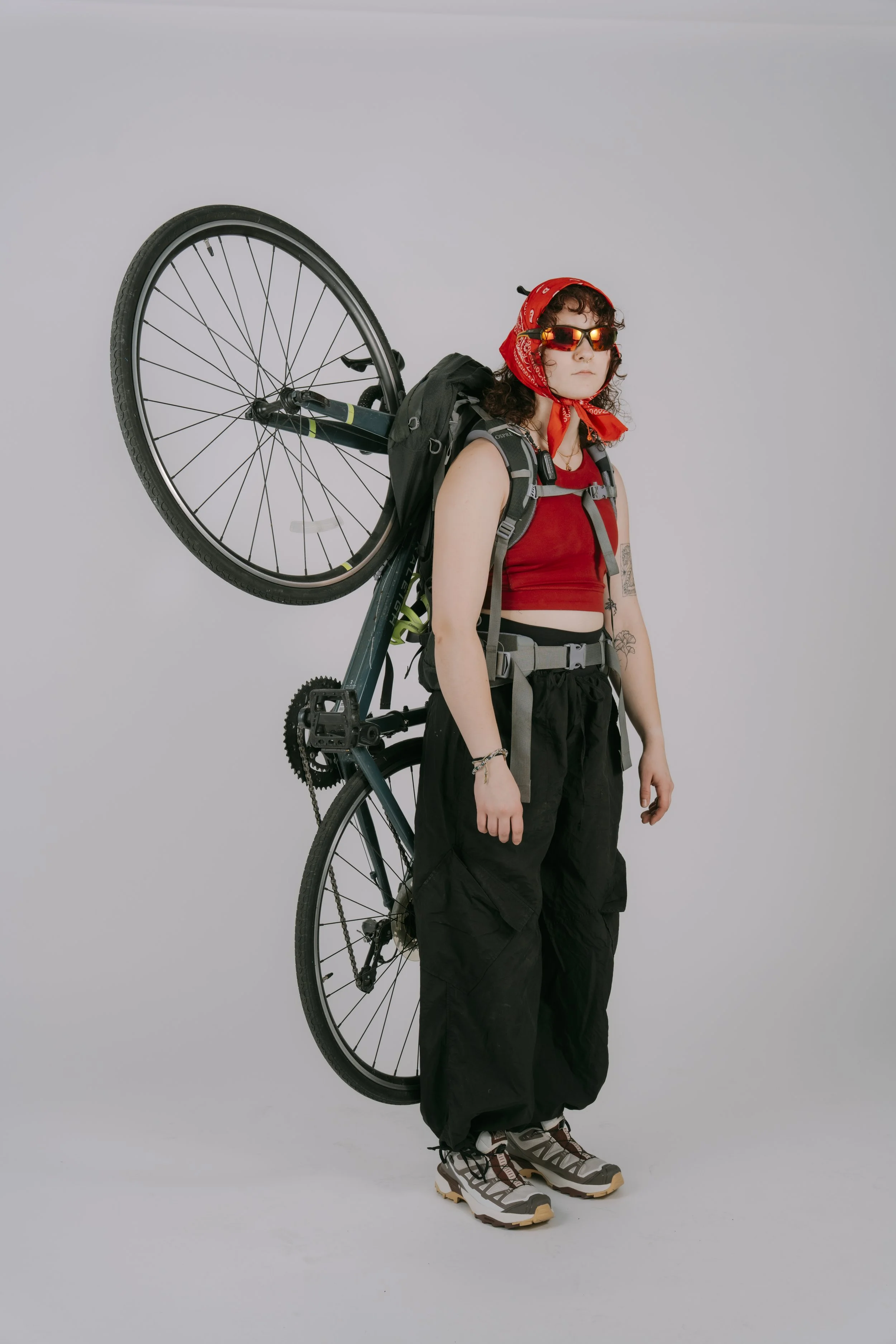 Woman dressed in outdoor gear with a bicycle on her back, wearing sunglasses, a red headscarf, and hiking shoes against a plain background.