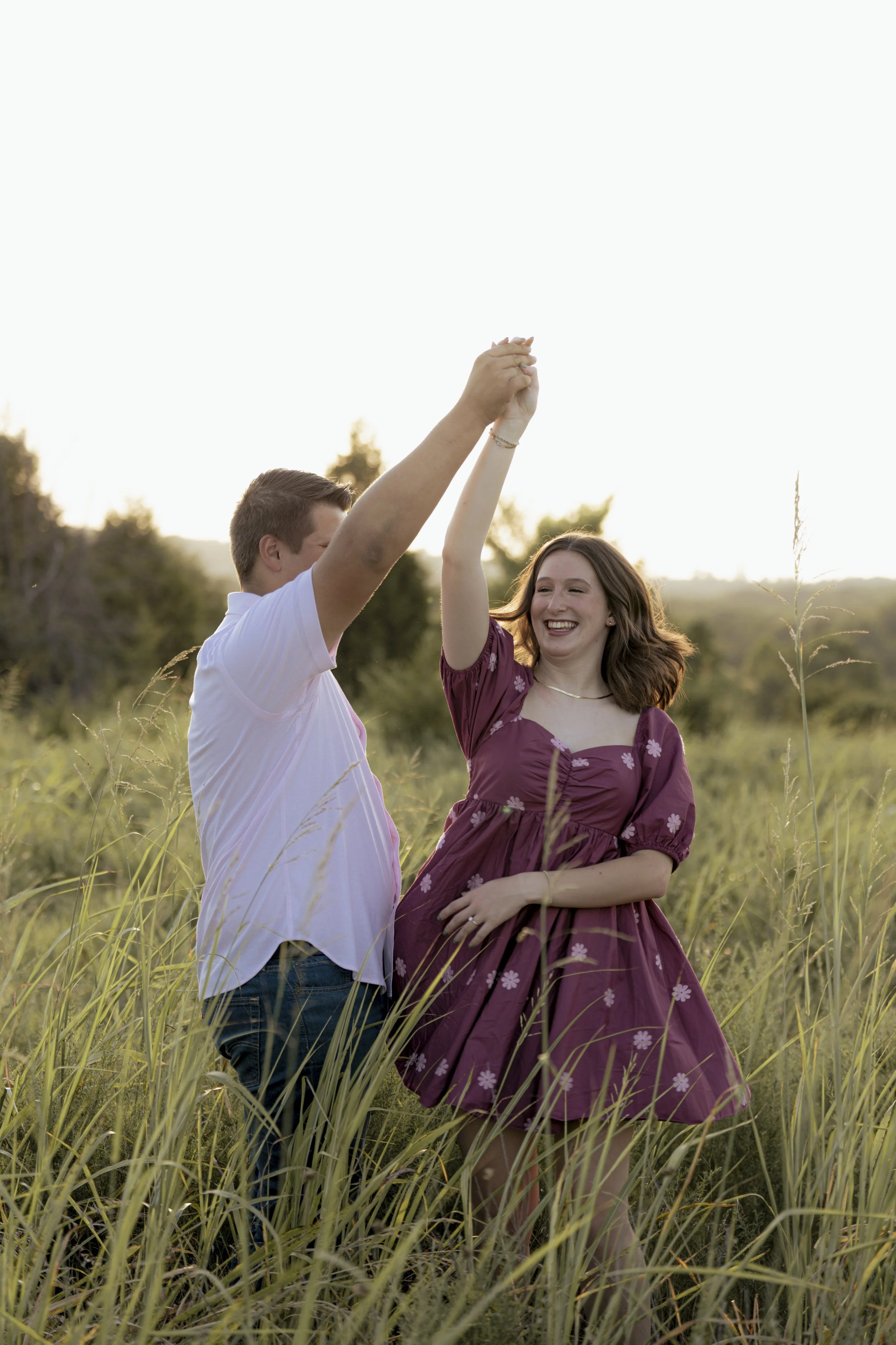 A young couple dancing and smiling in a grassy field at sunset, with the man in a white shirt and the woman in a purple dress.