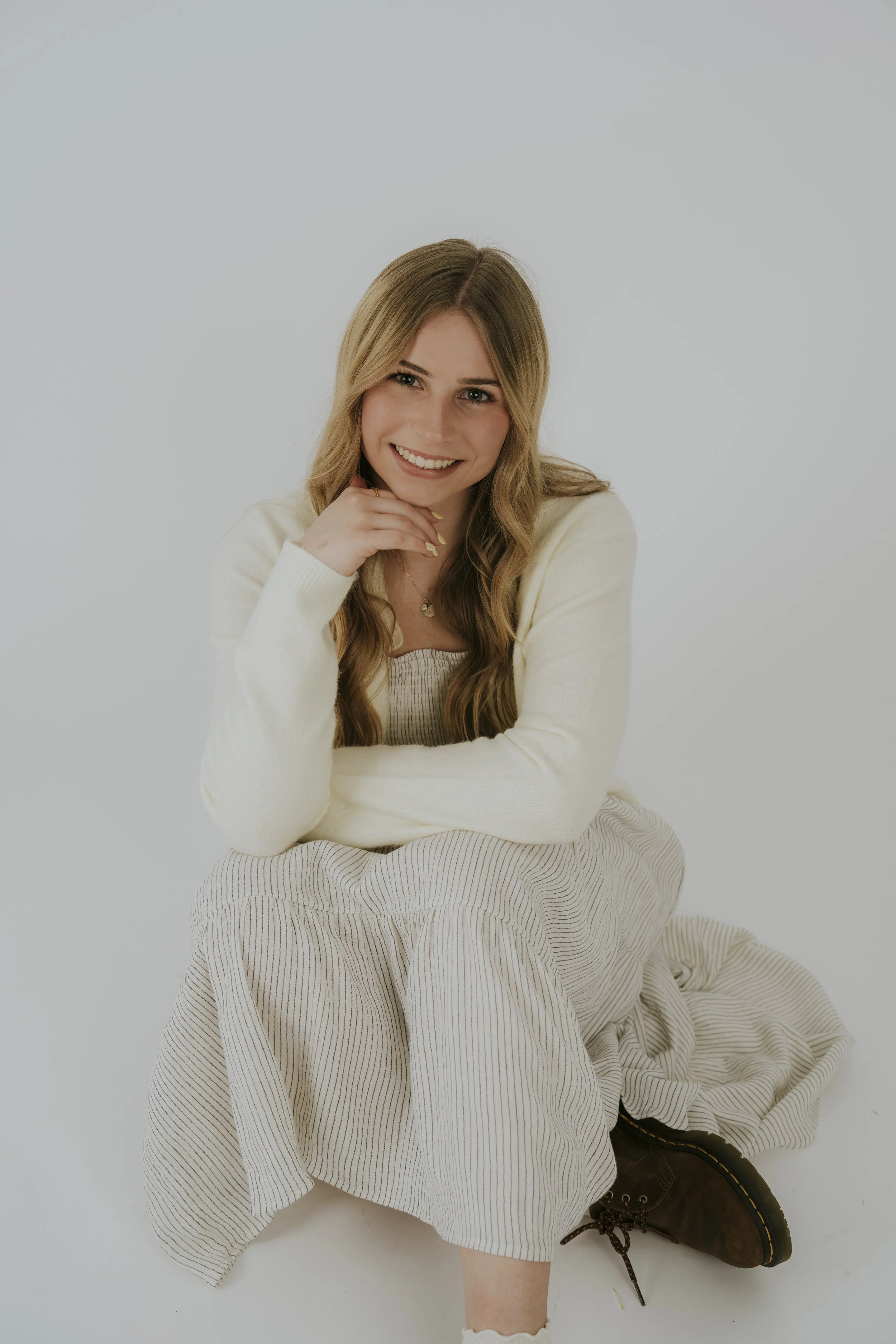 Young woman with long blonde hair, smiling, sitting on the floor with her legs crossed, wearing a cream sweater, a beige striped skirt, and brown boots, against a plain light background.