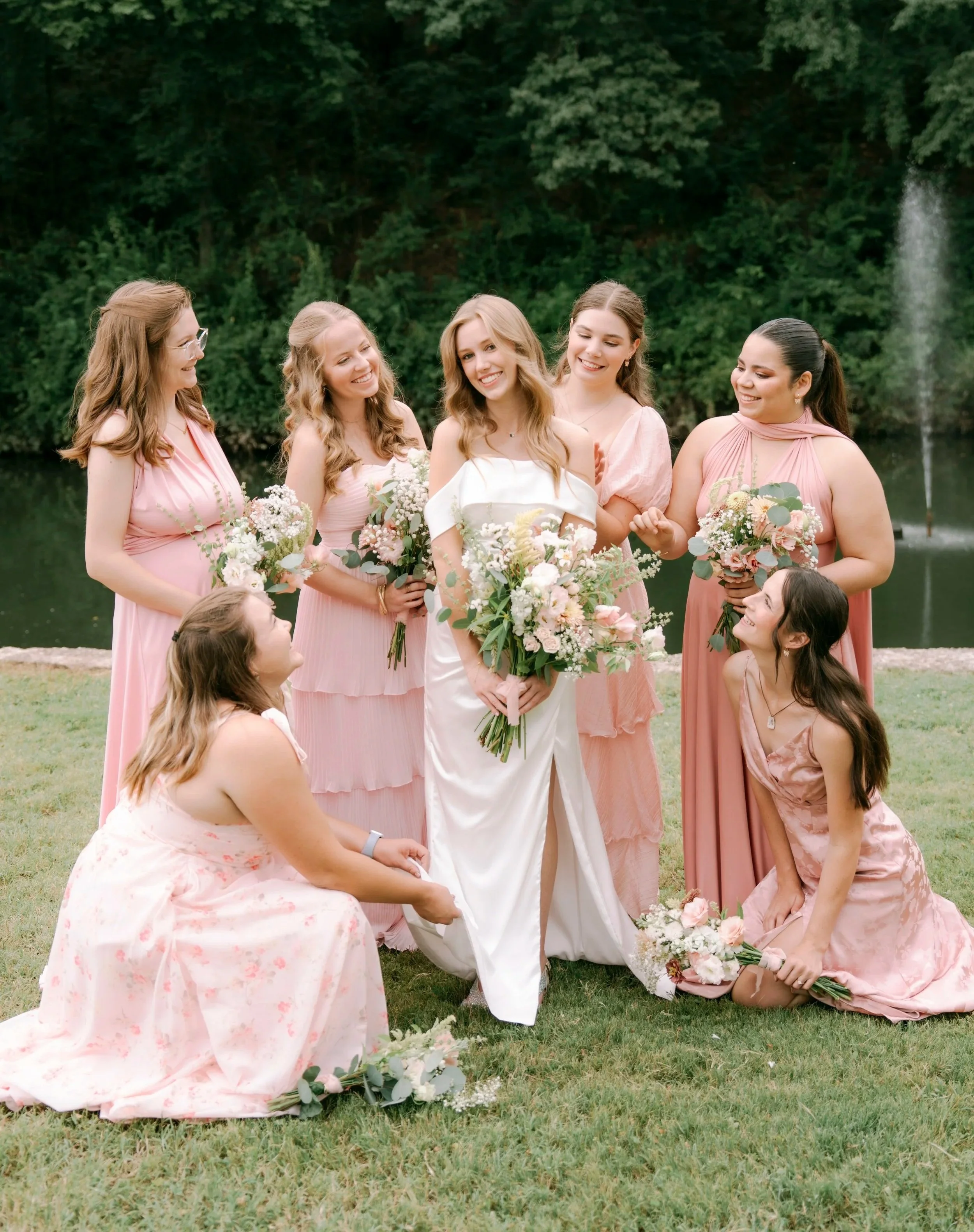 A bride and six bridesmaids outdoors near a fountain, all holding bouquets, with trees in the background.