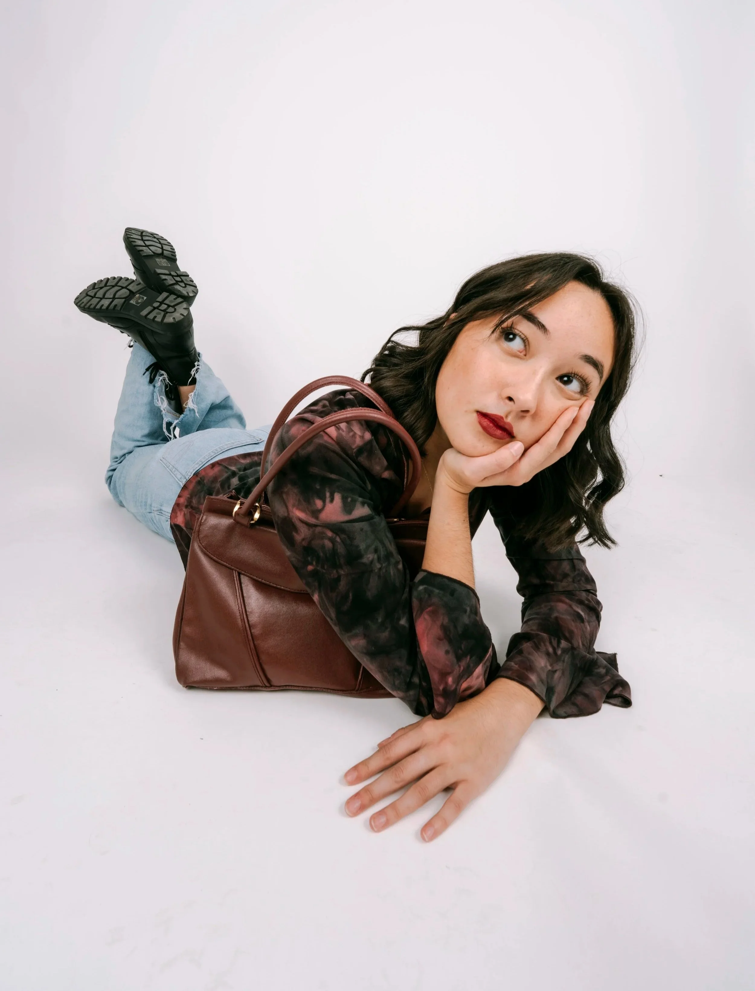 Young woman lying on her stomach on a white floor, resting her cheek on her hand, looking upward thoughtfully, with a brown handbag in front of her, wearing jeans, a dark patterned jacket, and black boots.