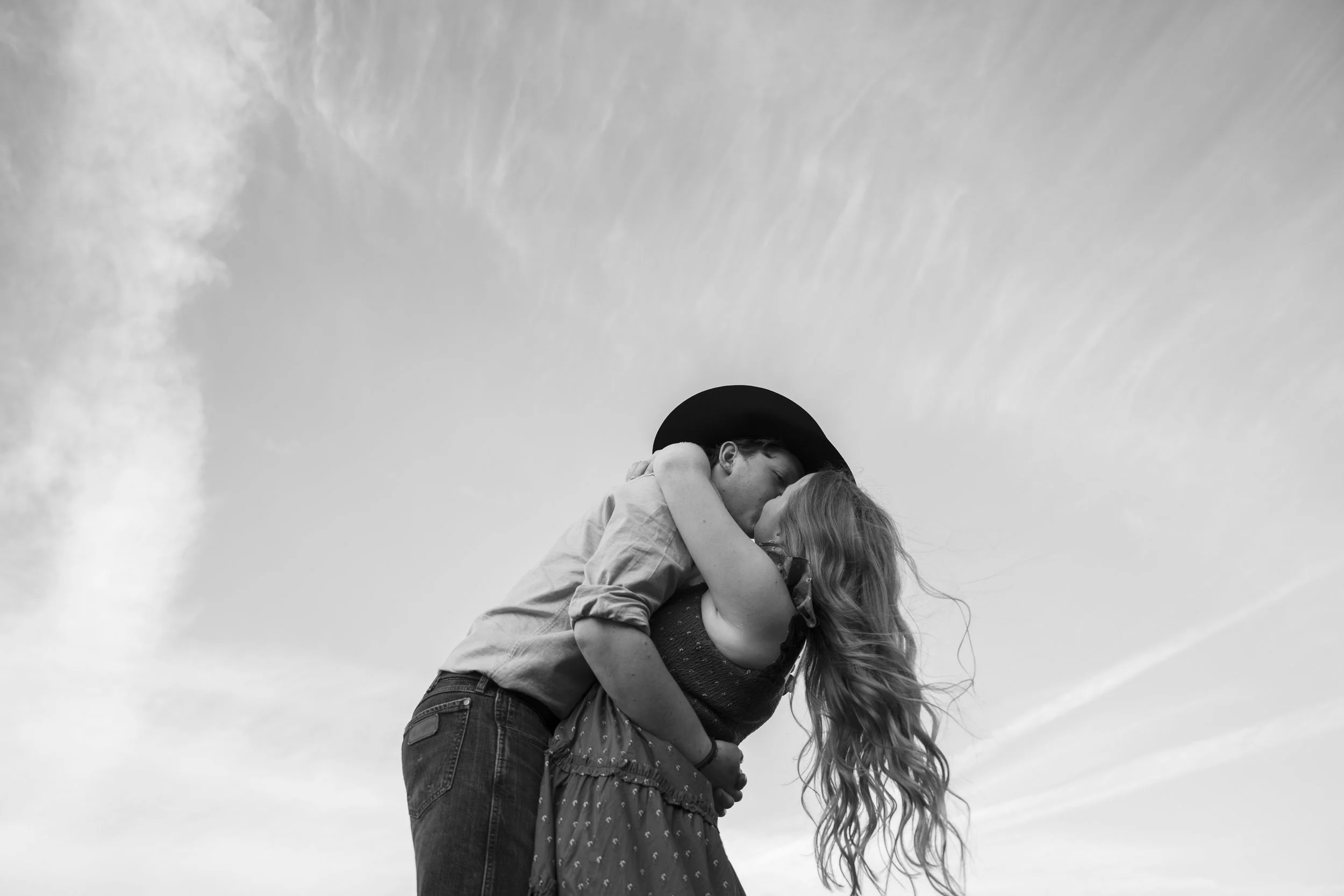 A black and white photo of a couple kissing, with the man wearing a wide-brimmed hat and the woman with long wavy hair, against a cloudy sky.
