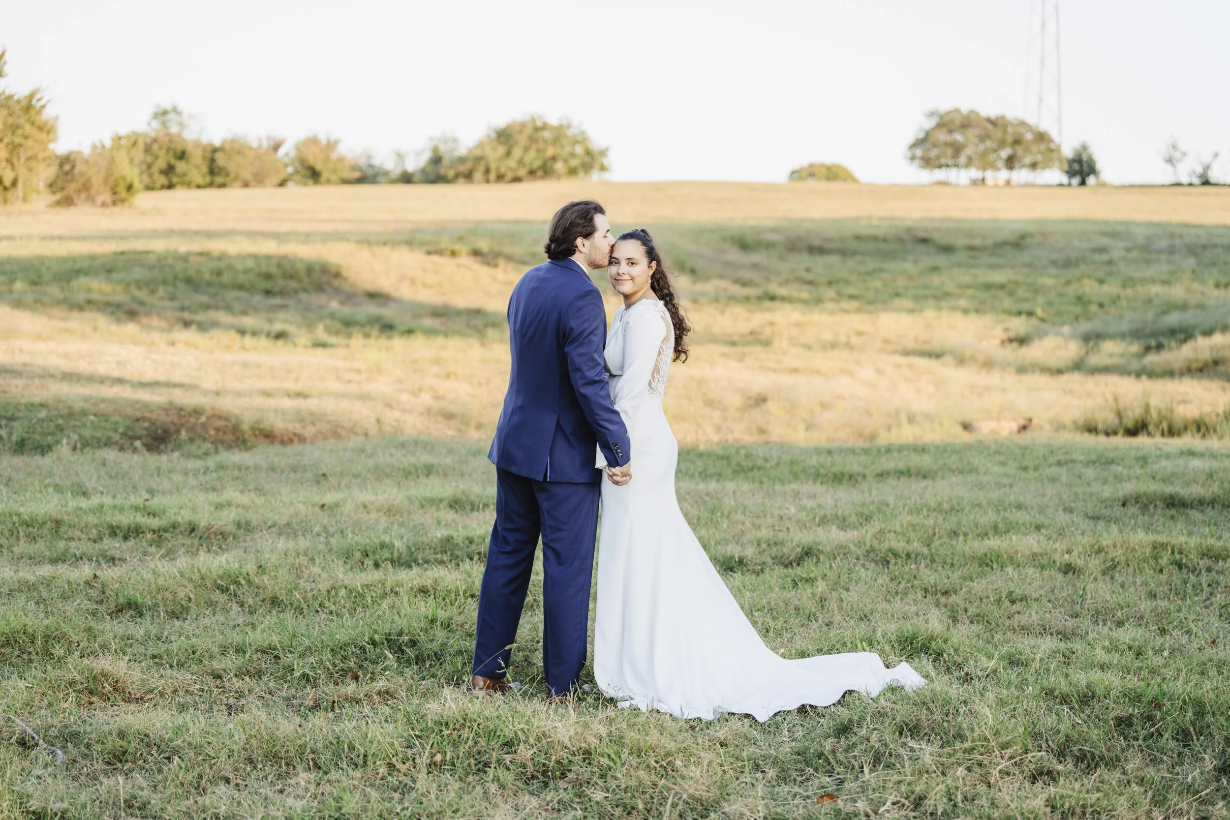 A newlywed couple holding hands in a grassy field, with rolling hills and trees in the background, during late afternoon.