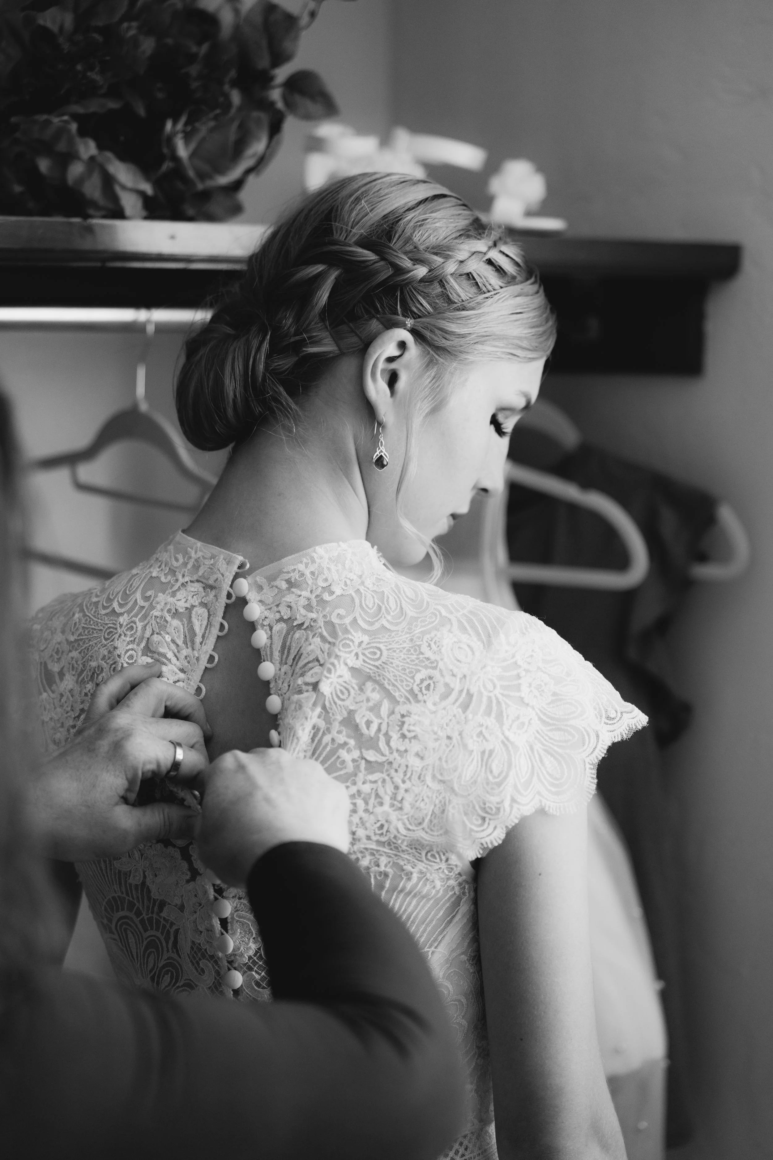 A woman in a lace dress is being assisted as she prepares for a special occasion, with someone fastening the buttons on her back.