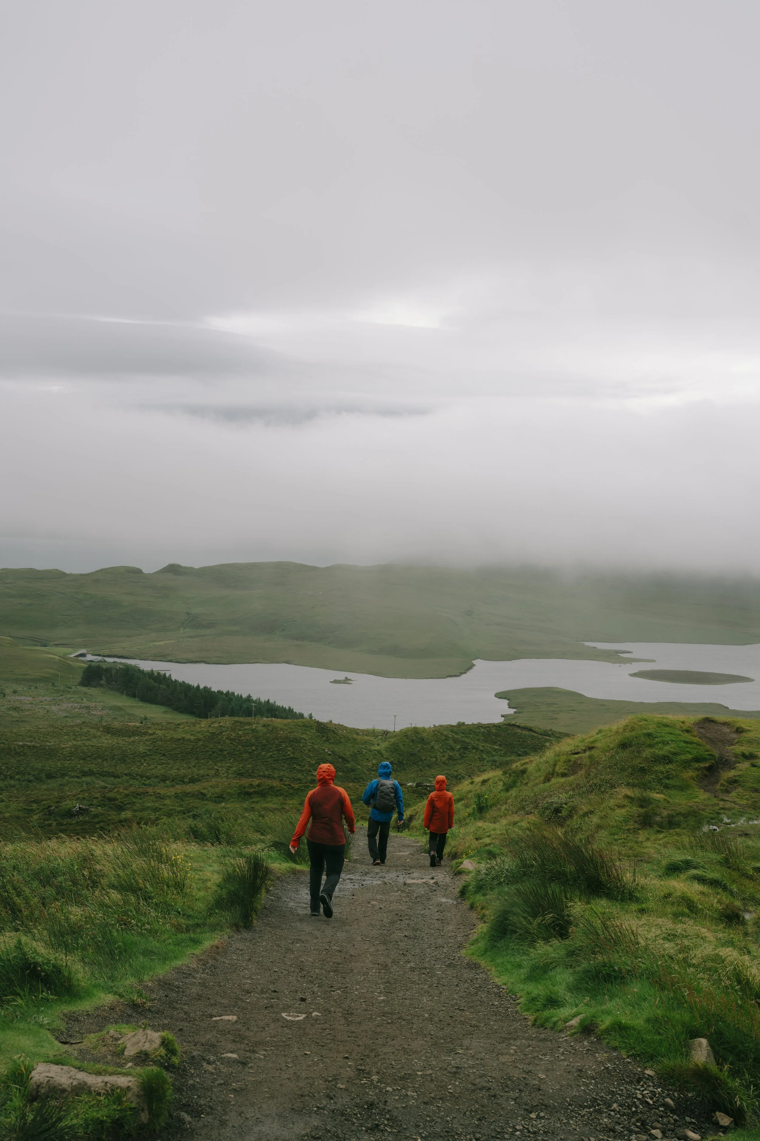 Three hikers walking down a dirt trail in green hilly landscape with a lake and cloudy sky in the background.