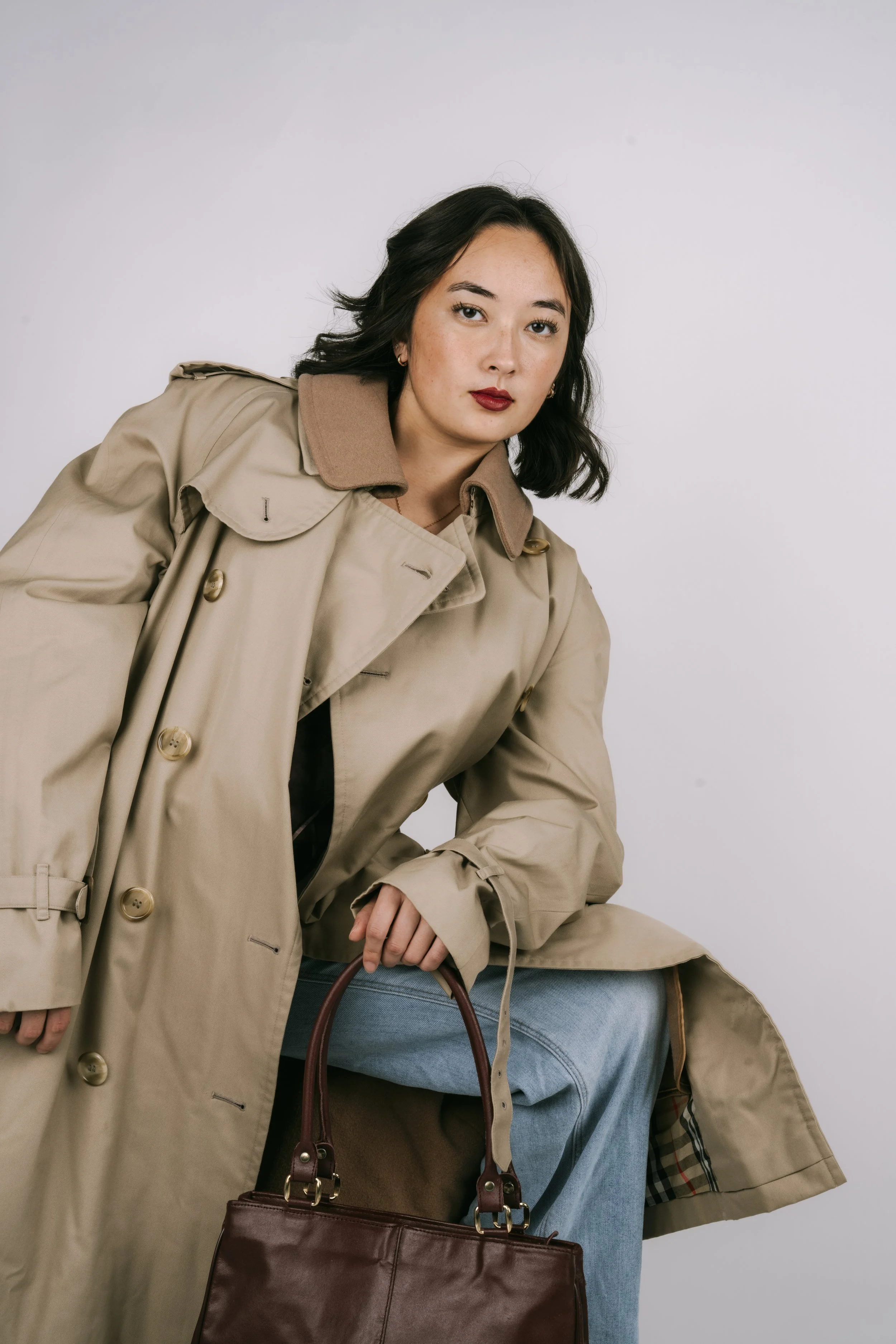 A woman with dark hair wearing a beige trench coat and blue jeans, holding a brown leather handbag, sitting against a plain white background.