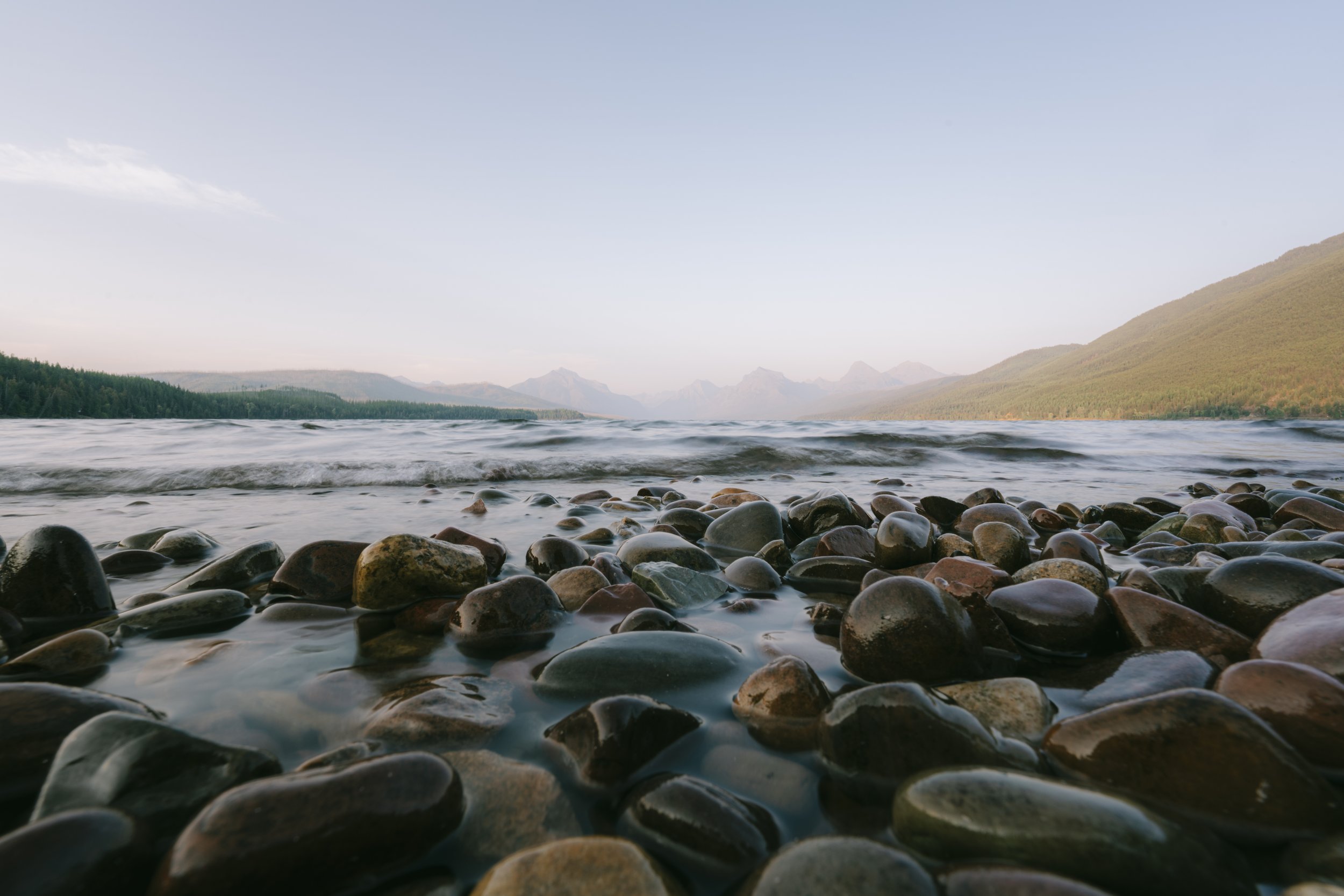 Pebble-covered shoreline with gentle waves, distant mountain range, and a clear sky.