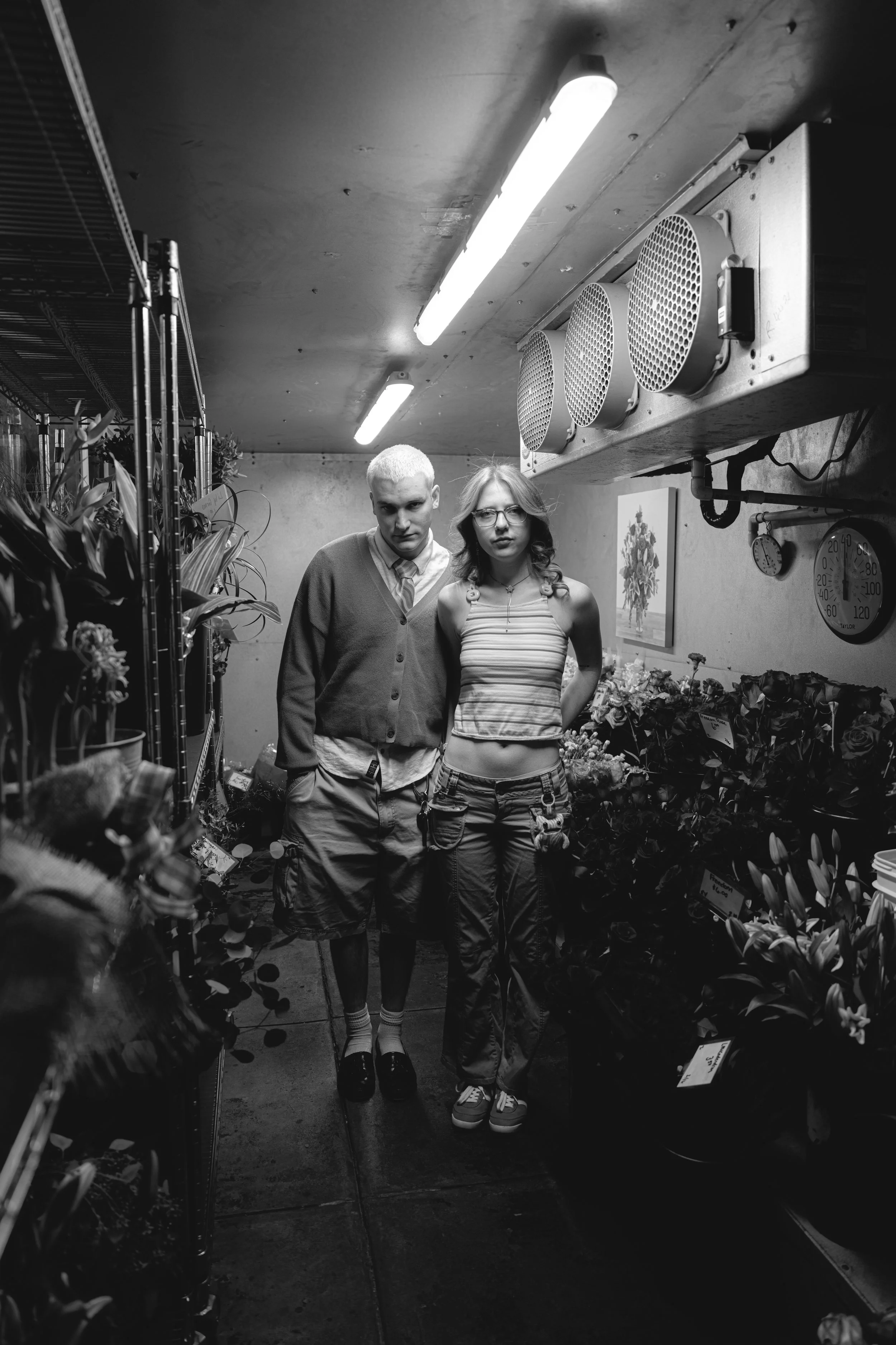 Two young people, a man and a woman, standing closely in a dark flower shop with shelves of plants and flowers on either side. Fluorescent lights are overhead, and there is a wall clock and wall art in the background.