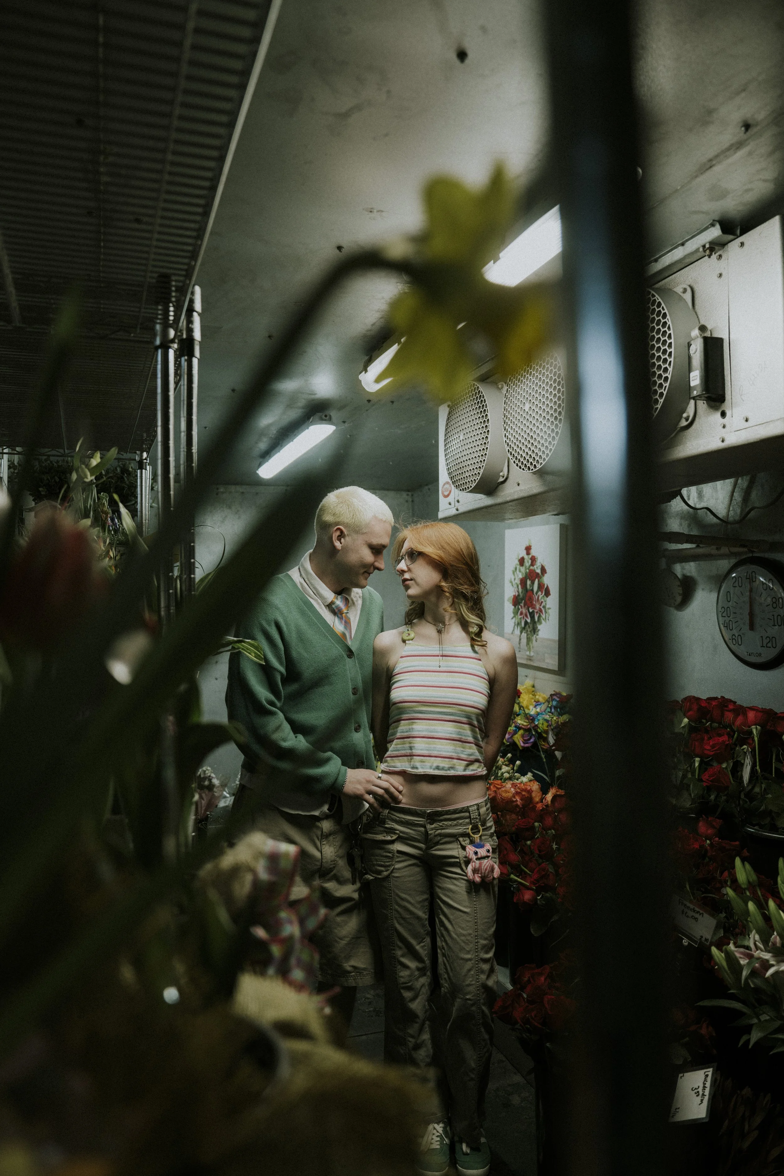 Two young people, a man with bleached blonde hair wearing a green cardigan and a woman with red hair wearing a striped crop top, standing close together inside a flower shop surrounded by various flowers and plants, with artificial lighting overhead.