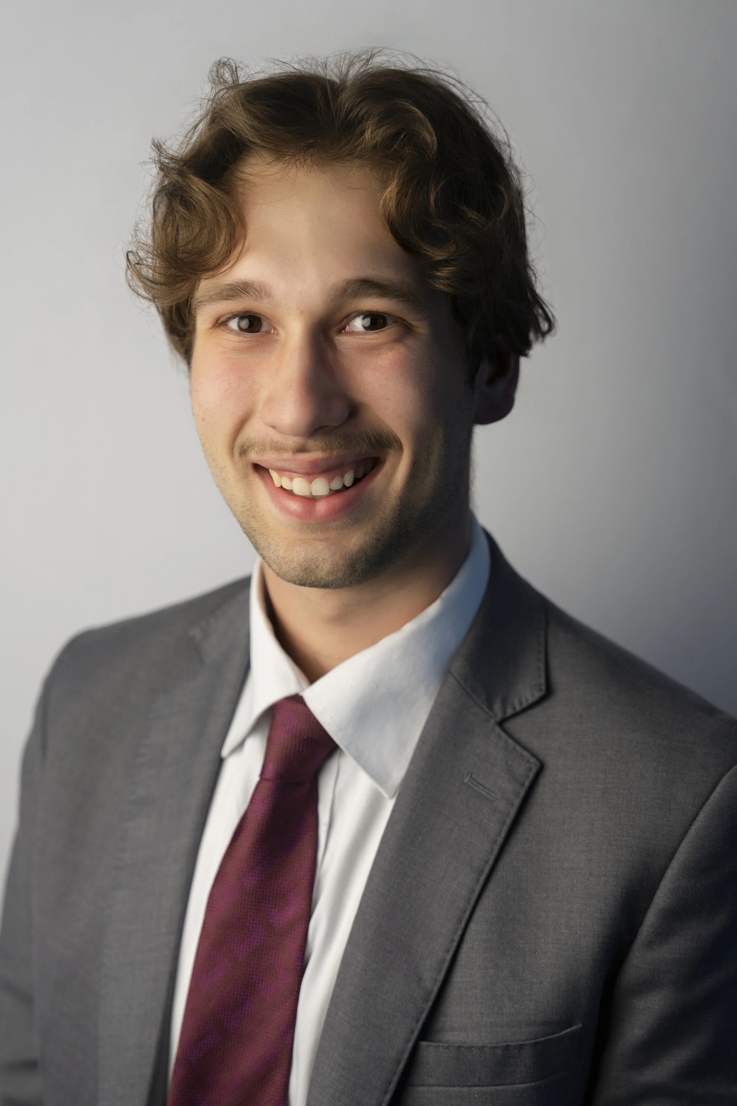 Portrait of a young Caucasian man in a gray suit, white shirt, and burgundy tie, smiling against a plain light gray background.