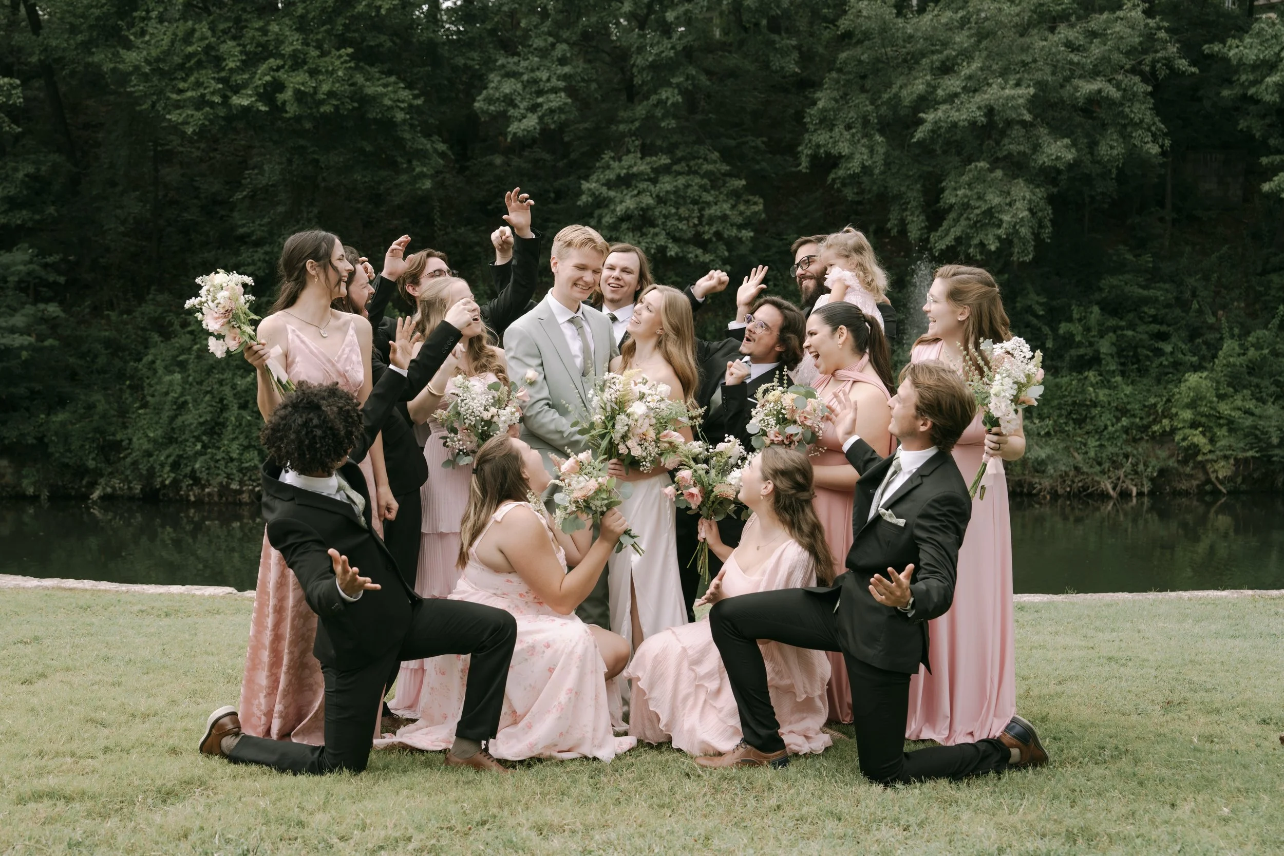 A large wedding party outdoors near a body of water, celebrating and smiling, with women holding bouquets and men in tuxedos.