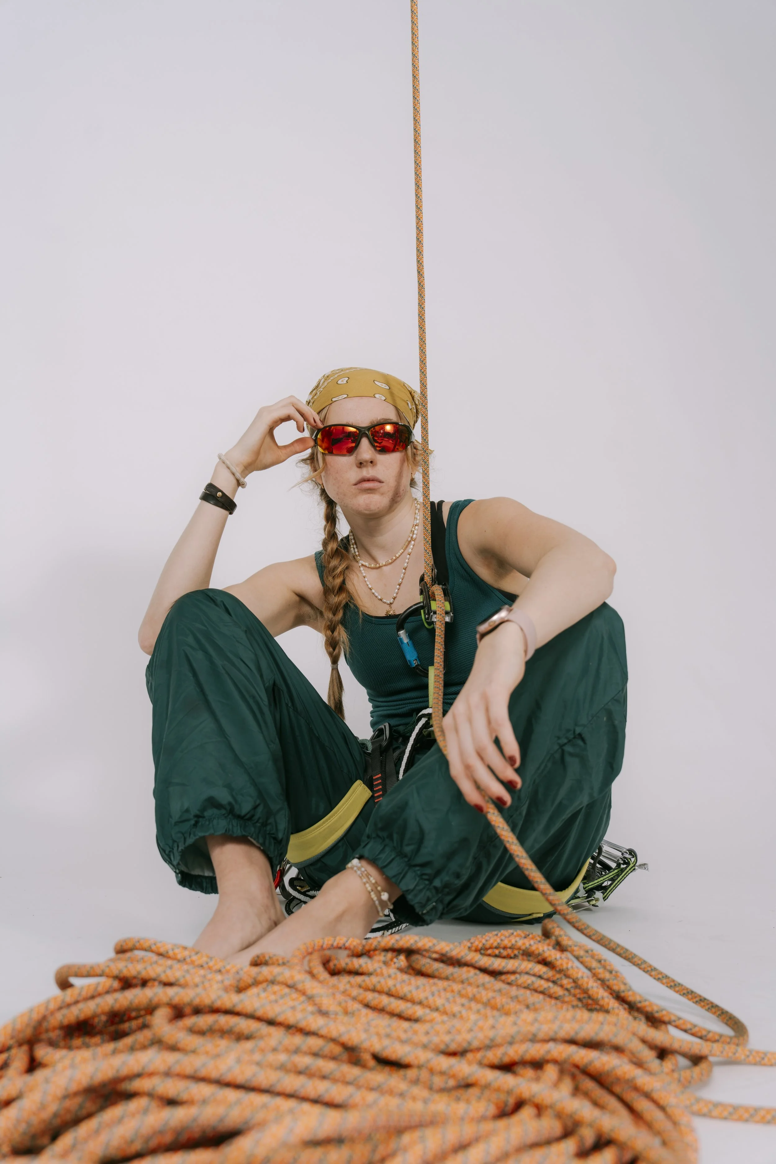 A woman dressed in rock climbing gear, sitting on the floor with a large coiled climbing rope in front of her. She is wearing sunglasses, a bandana, necklaces, and bracelets, with a focused expression and a harness attached to the rope.