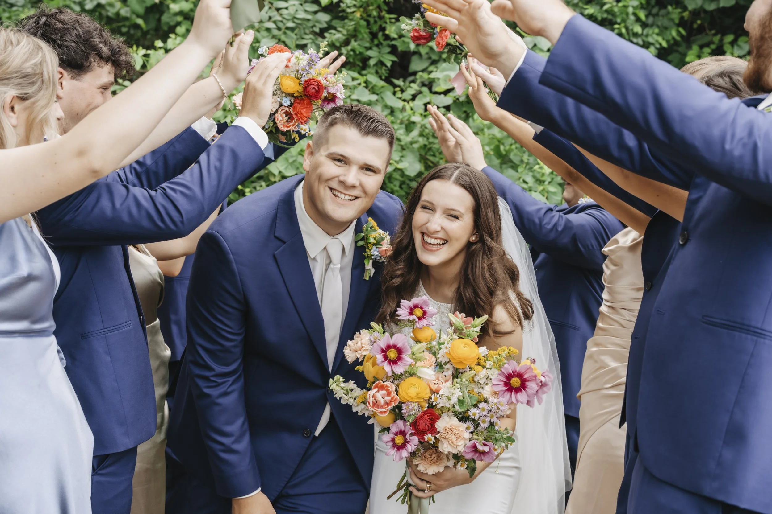 A happy bride and groom smiling under a flower arch made by their wedding guests in a lush green outdoor setting.