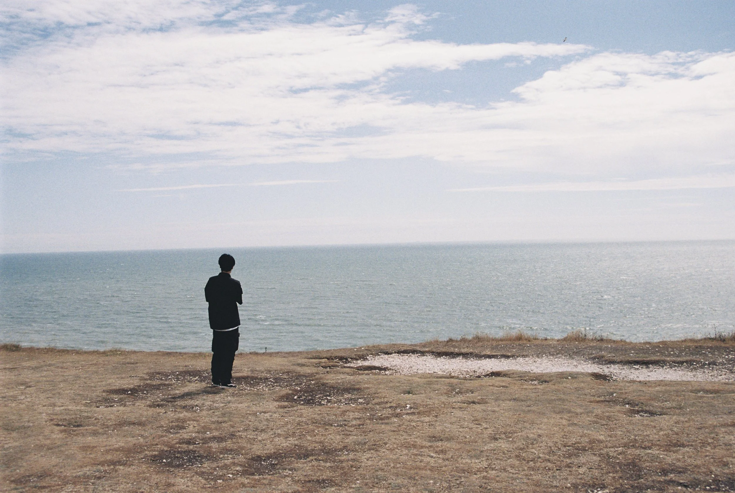 A person standing alone on a dirt path, looking out at the ocean with clouds in the sky.