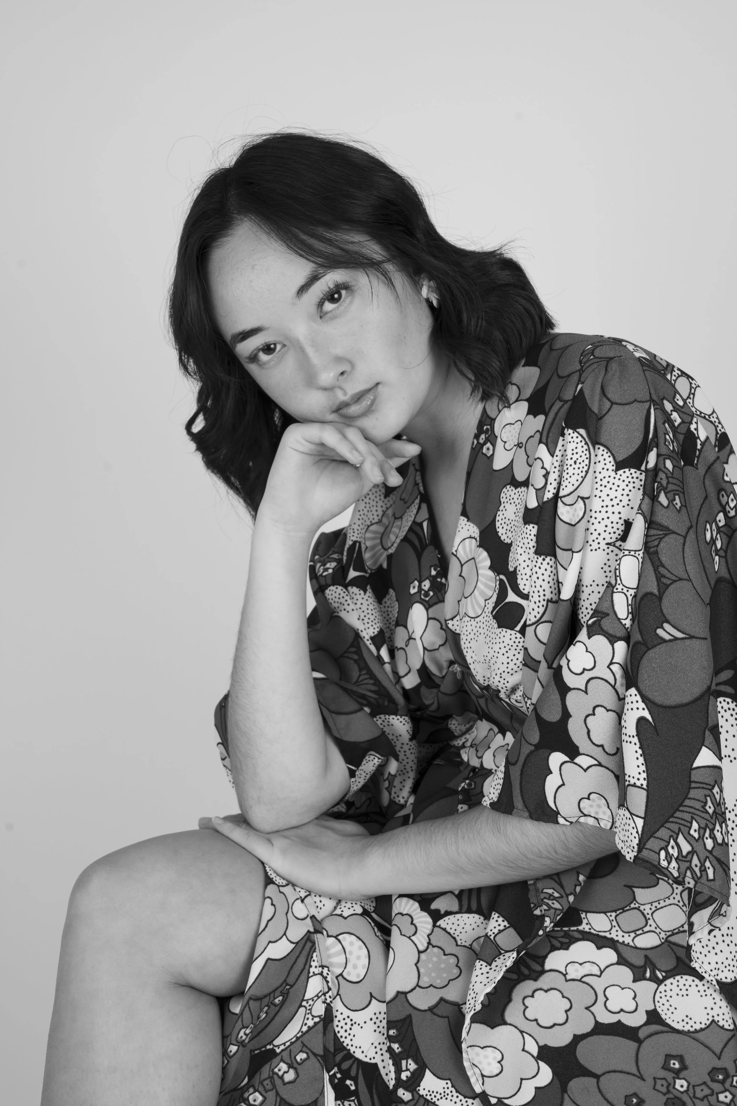 Black and white portrait of a woman sitting and resting her chin on her hand, wearing a patterned floral kimono with short dark hair and earrings.