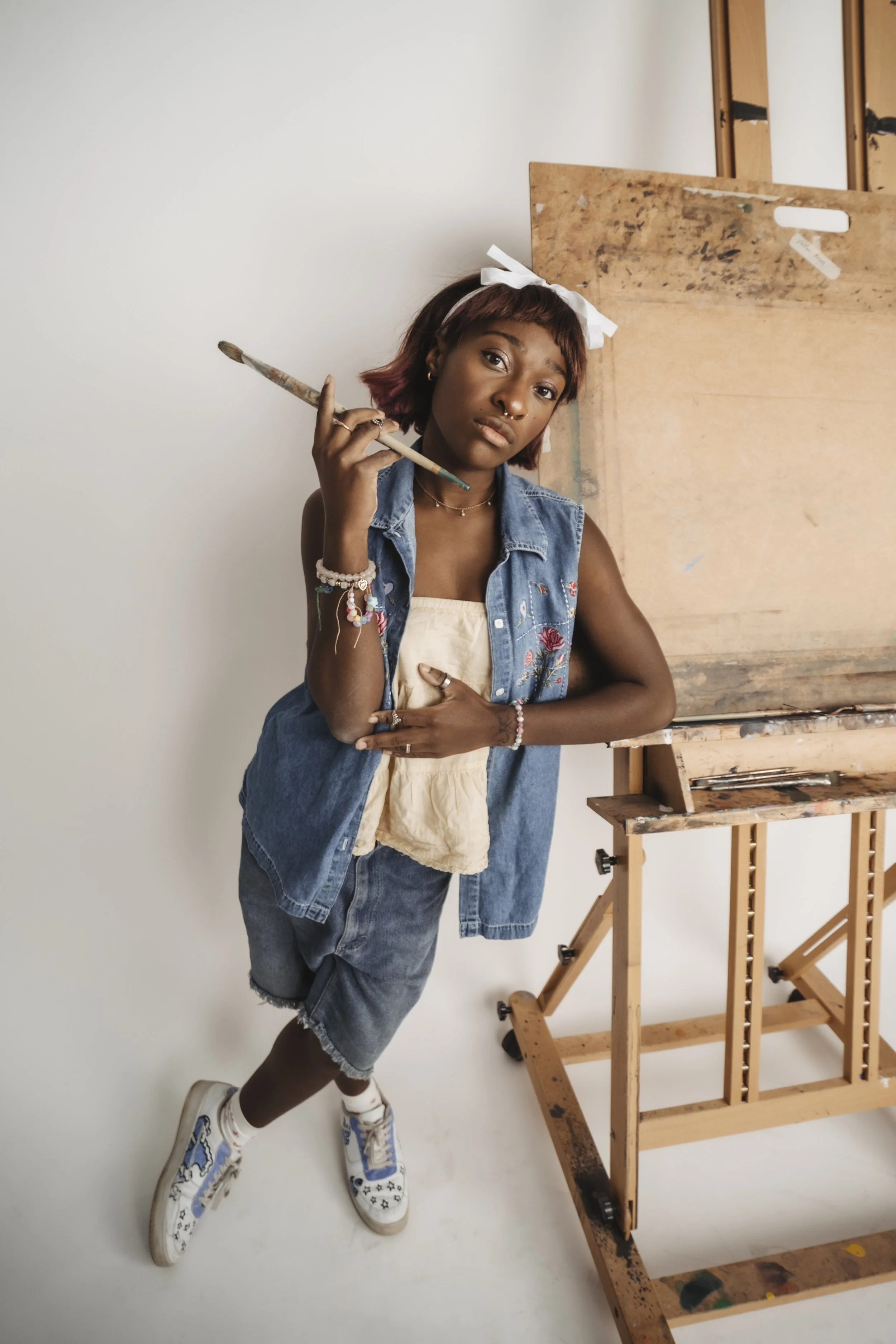 A young woman standing in an art studio, holding a paintbrush near her head, wearing casual denim clothing, jewelry, and a white headband with a bow, in front of a wooden easel and blank canvas.