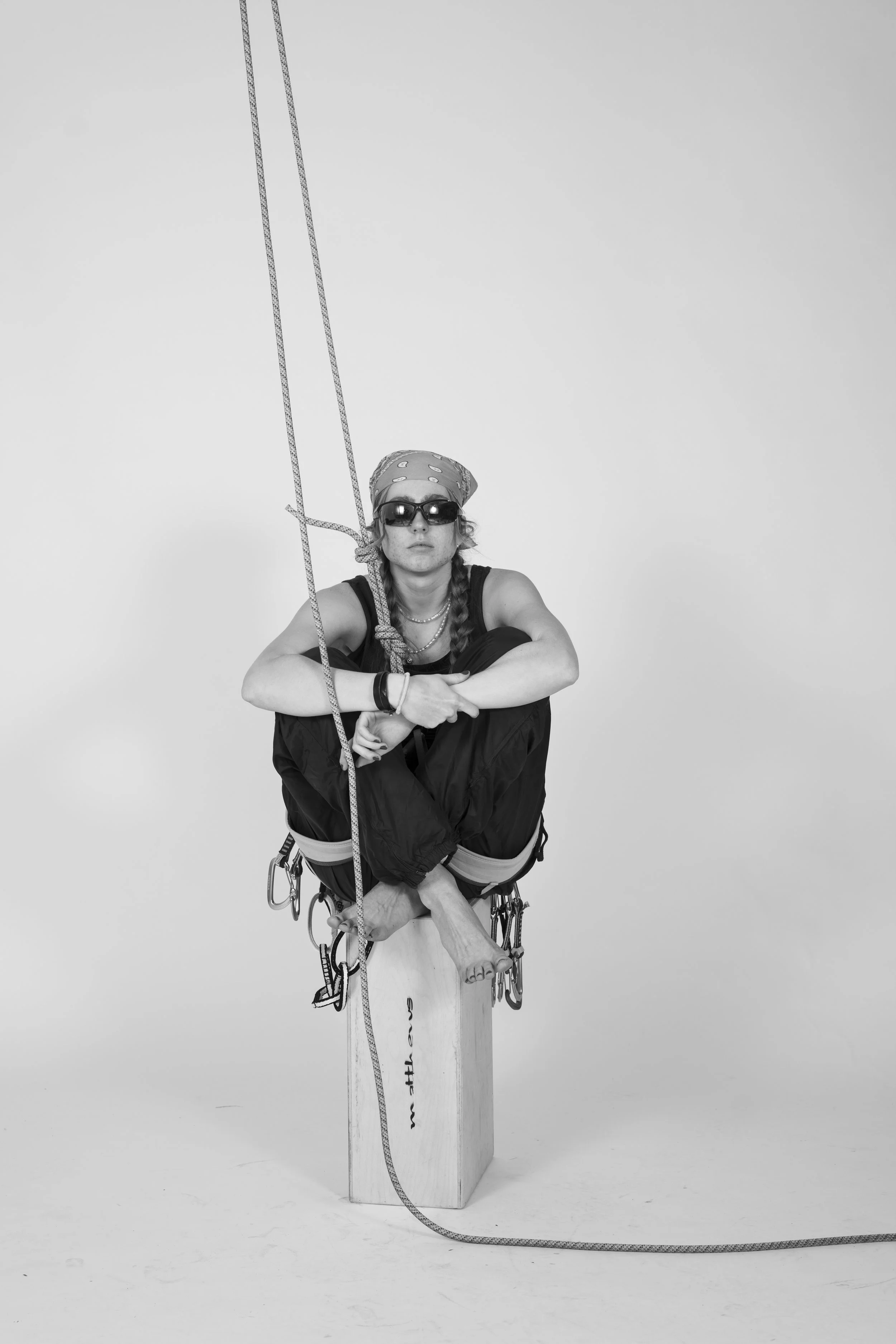 A woman with sunglasses, a bandana, and jewelry sits on a wooden block, holding a rope attached to a setup resembling a swing, in a studio with a plain background.