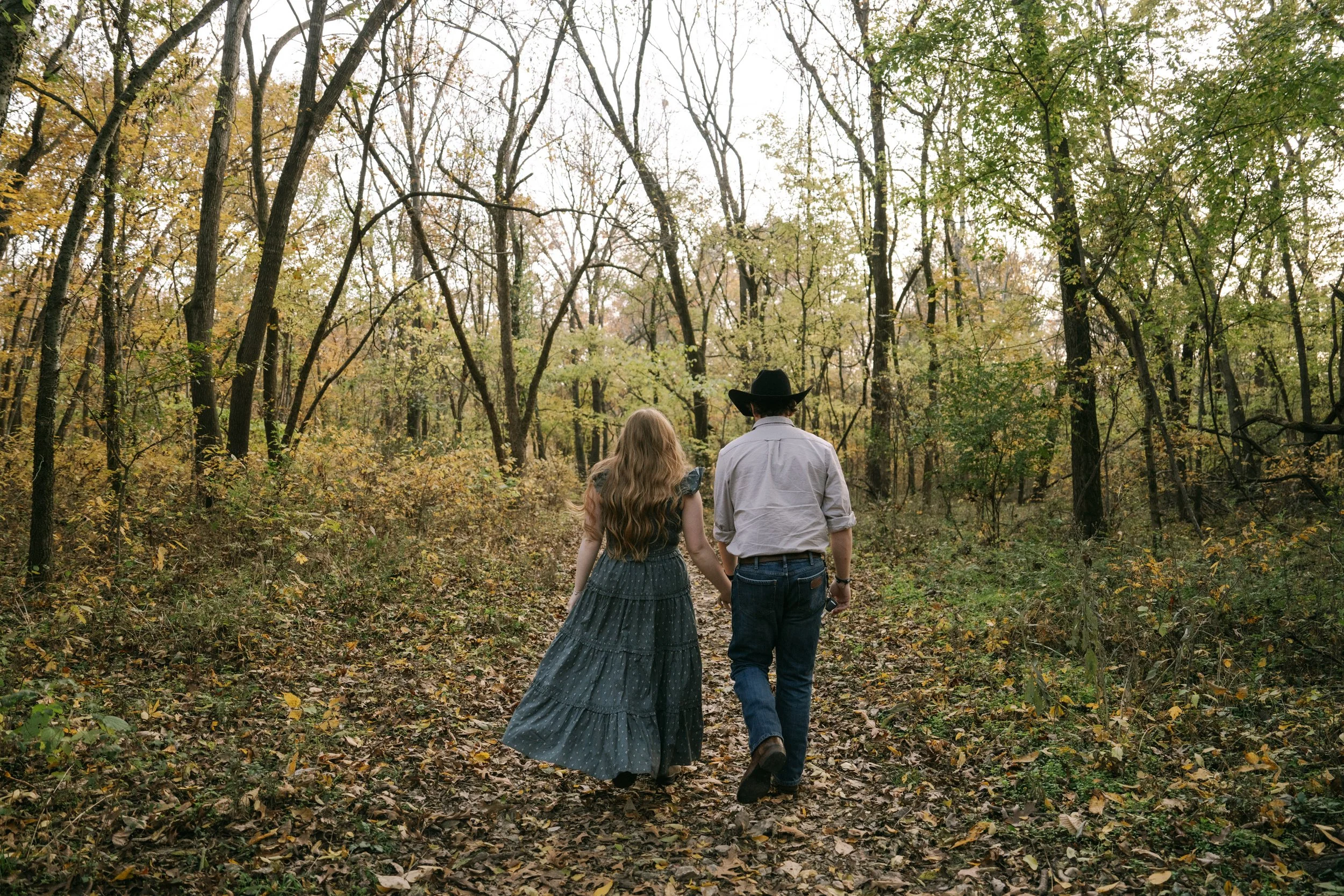A couple walking hand in hand through a wooded forest in autumn, with trees with changing leaves and a leaf-covered trail.