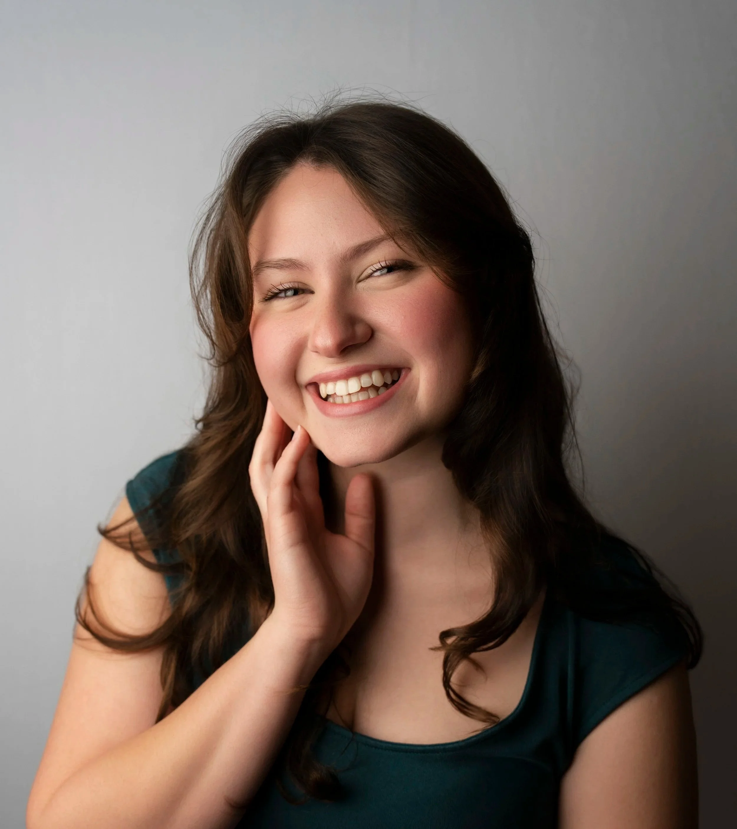 A young woman with long brown hair, smiling and gently touching her face with her hand, wearing a teal top, against a neutral background.