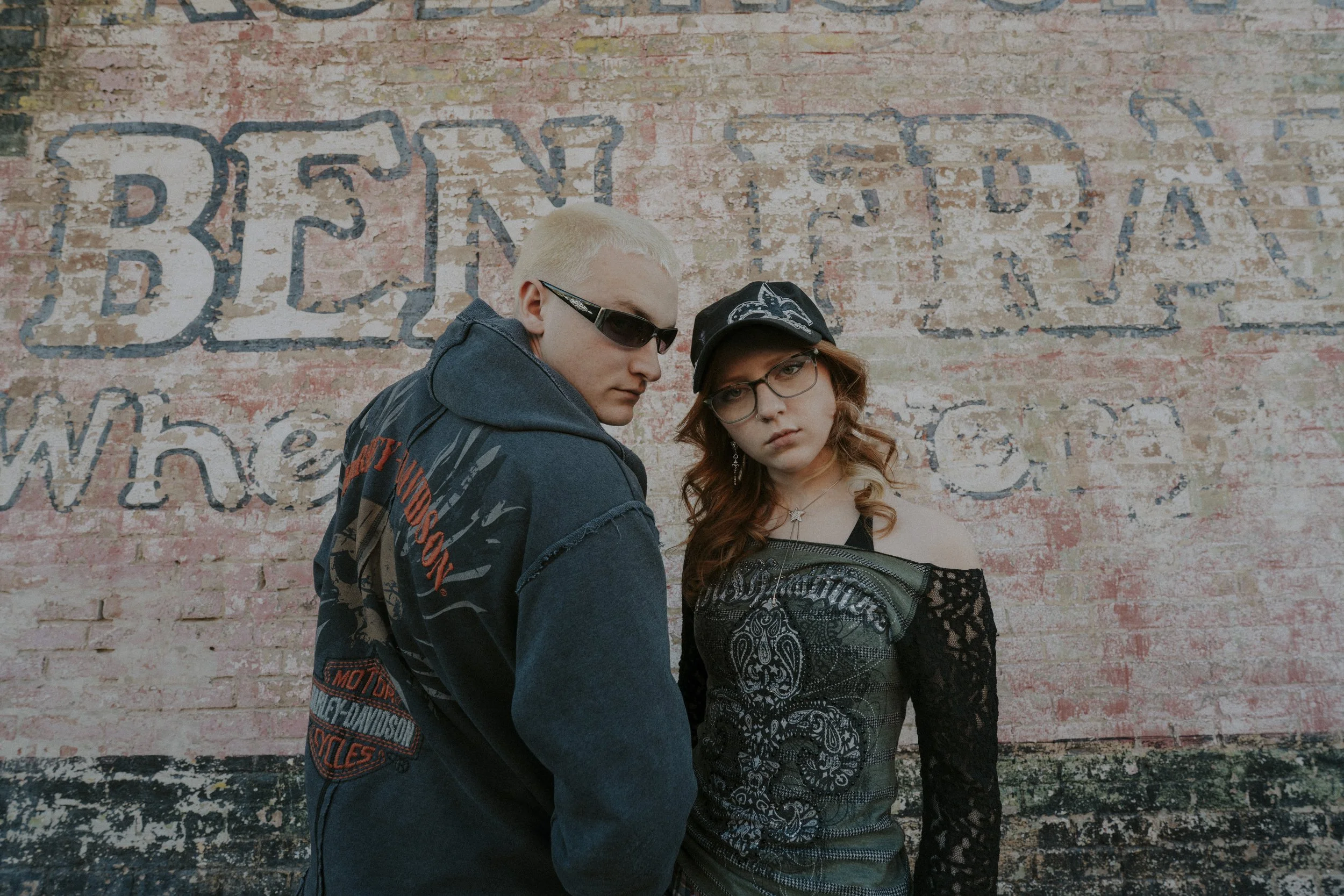 A young man with blond hair and sunglasses standing next to a young woman with red hair, glasses, and a black cap, both dressed in dark, edgy clothing, posing against a weathered brick wall with faded painted text.