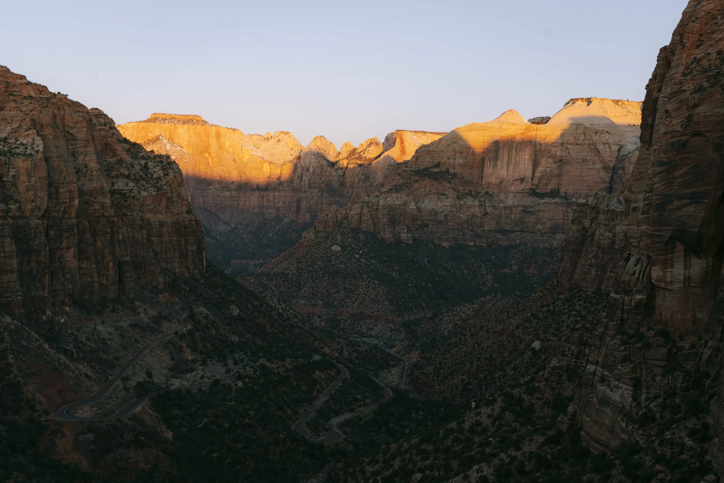 Sunset illuminating the tops of canyon walls in Zion National Park, with winding roads visible in the dark valley below.