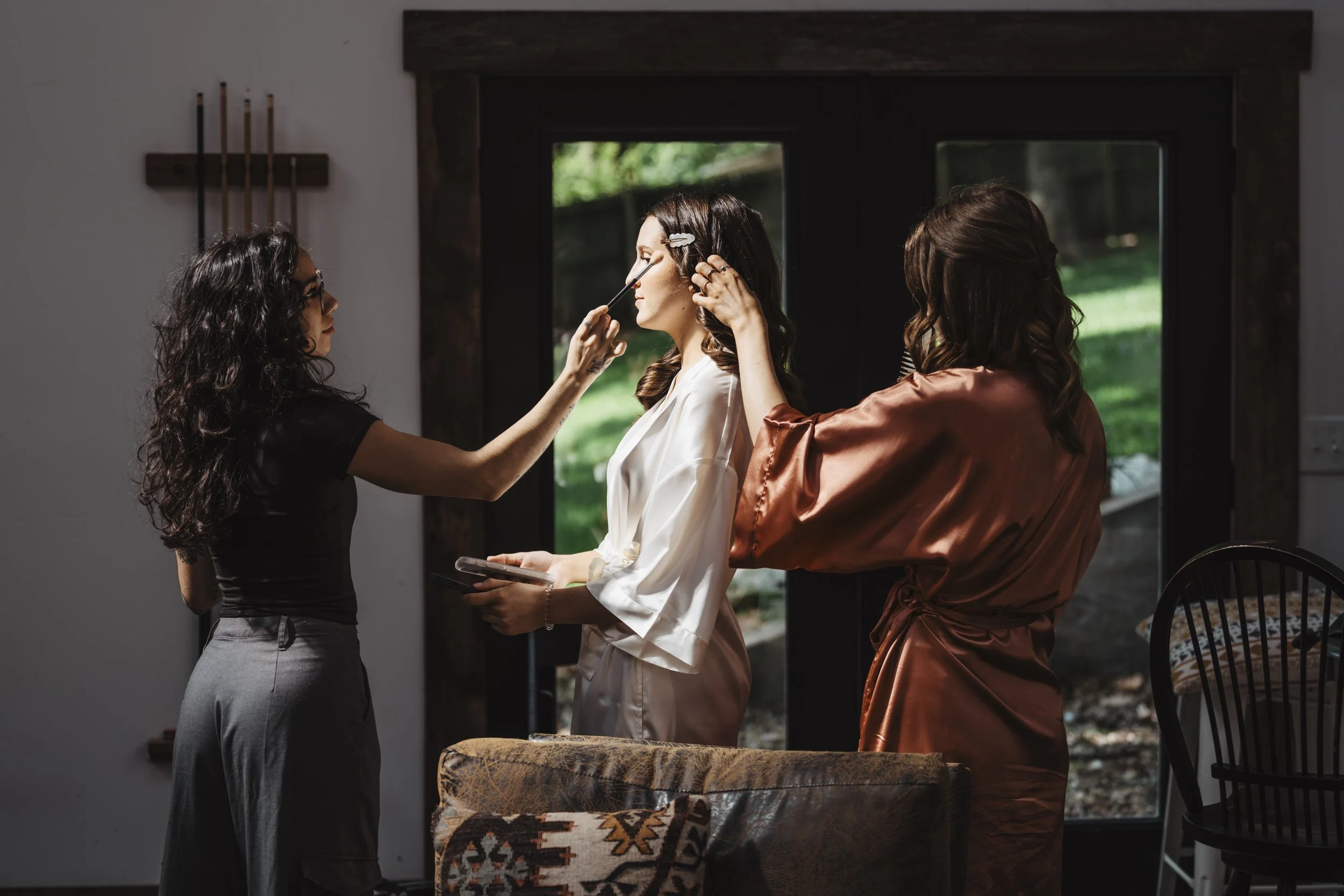 A woman in a white robe is getting her makeup done by a makeup artist, while another woman in a satin robe styles her hair, inside a room with a large window showing greenery outside.