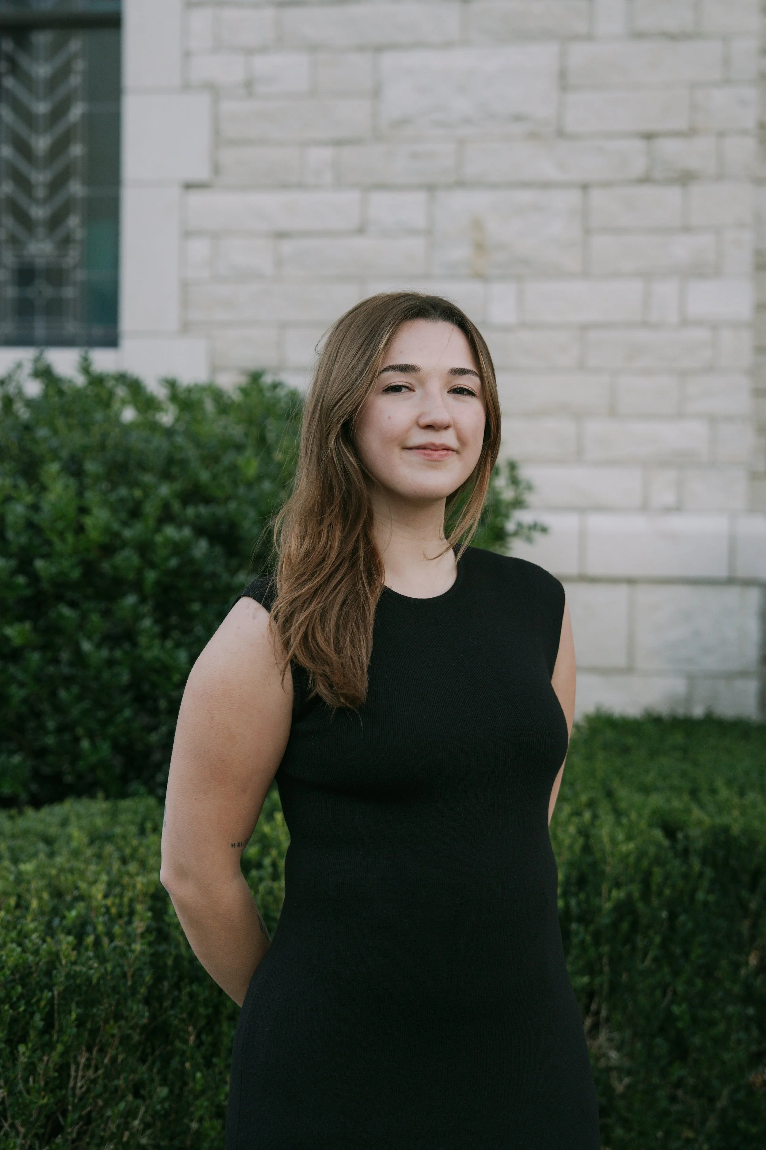 A young woman with brown hair wearing a sleeveless black dress standing outdoors in front of green bushes and a stone building.