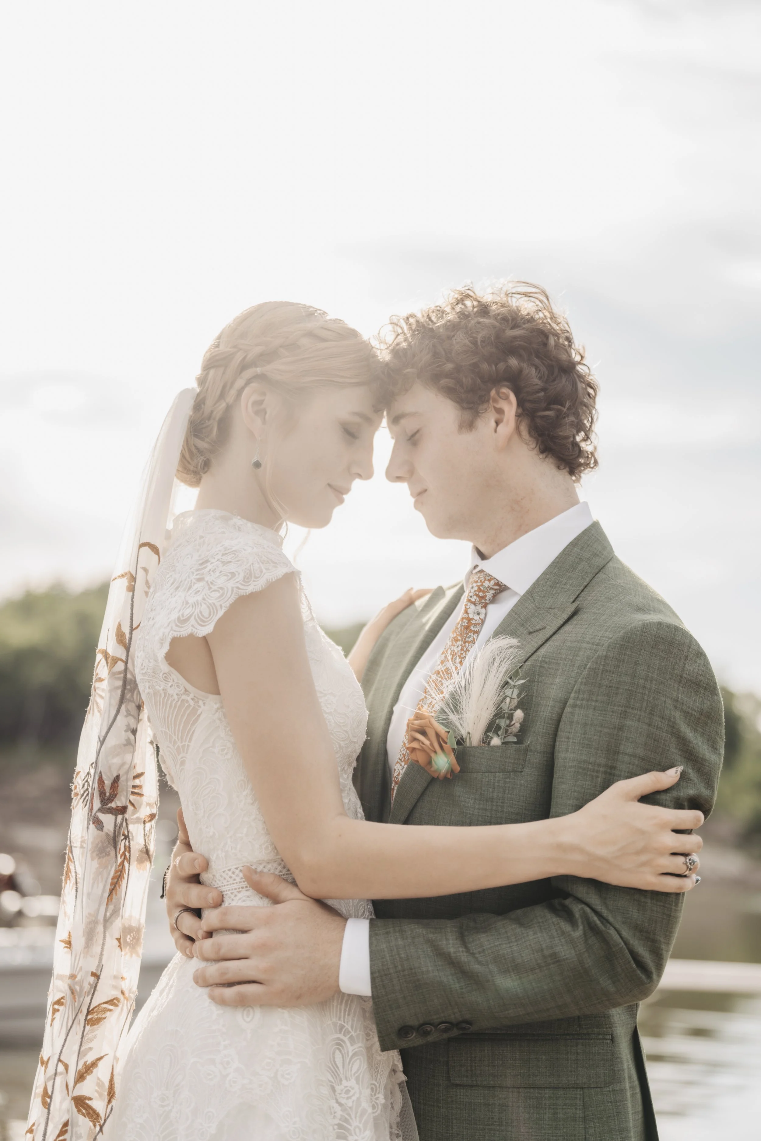 A bride and groom embrace outdoors with forehead and nose touching, eyes closed, during their wedding.