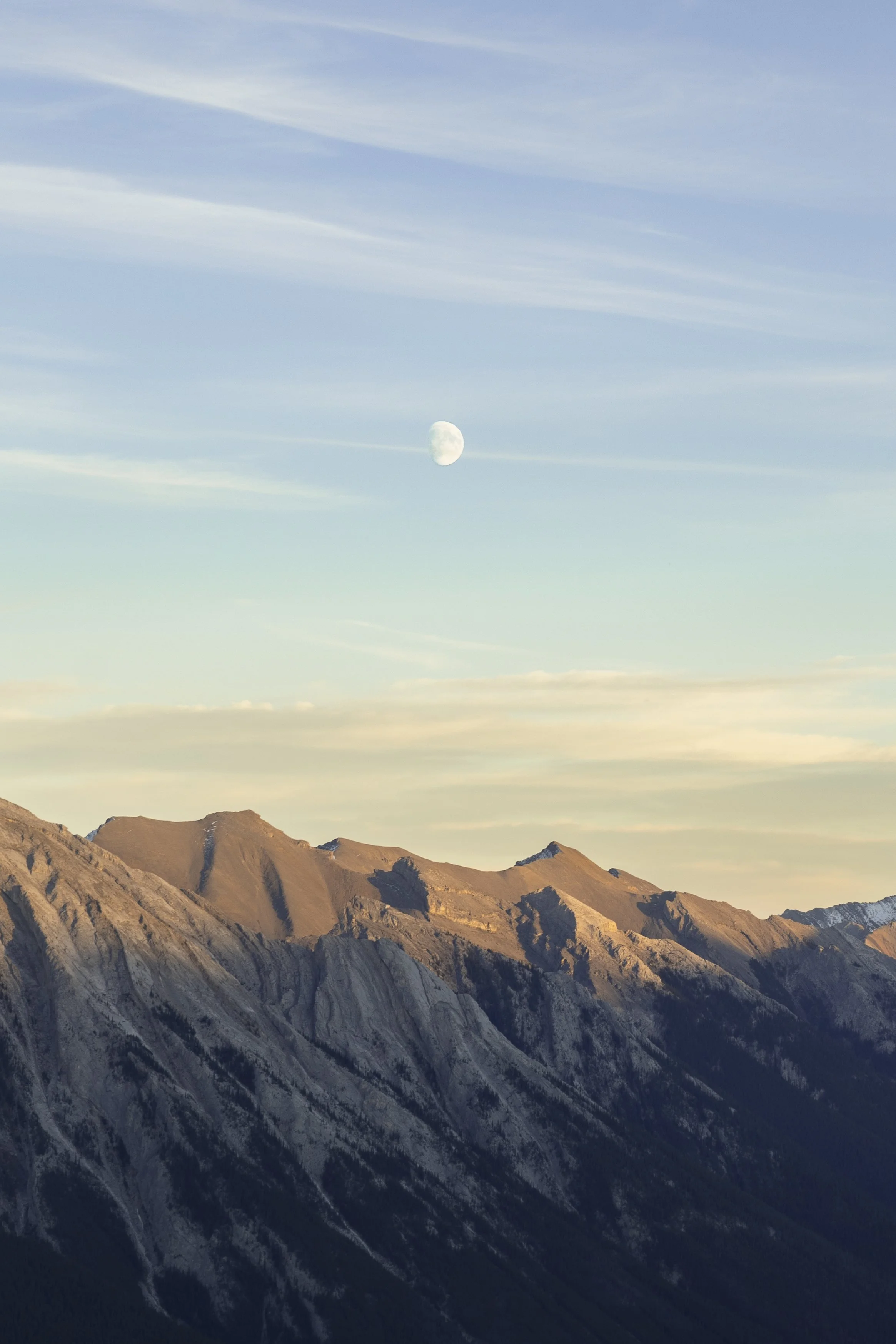 Mountain range under a blue sky with a visible moon and wispy clouds.