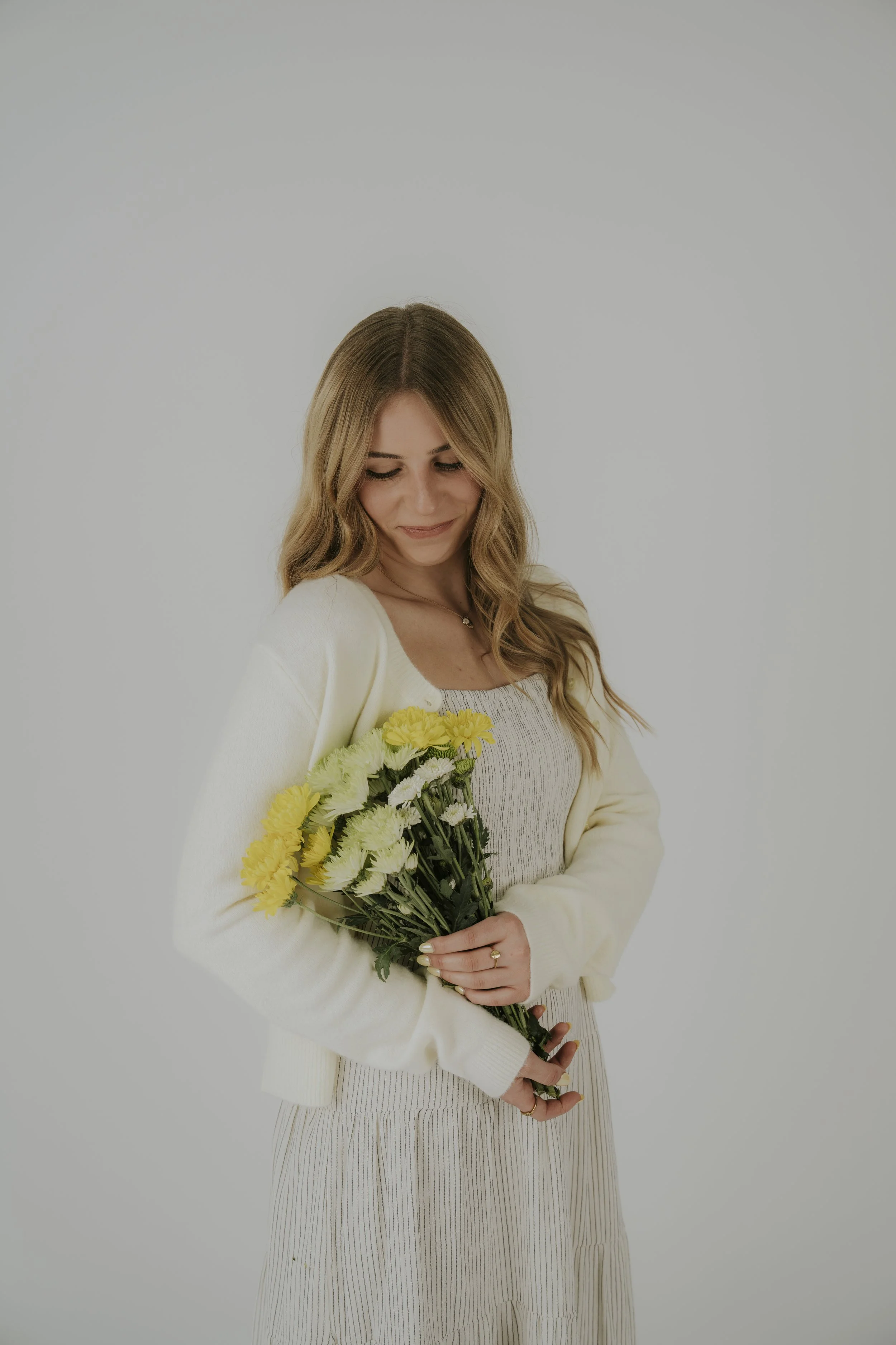 A young woman with long wavy blonde hair holding a bouquet of yellow and white flowers, wearing a light-colored dress and cream cardigan, against a plain white background.