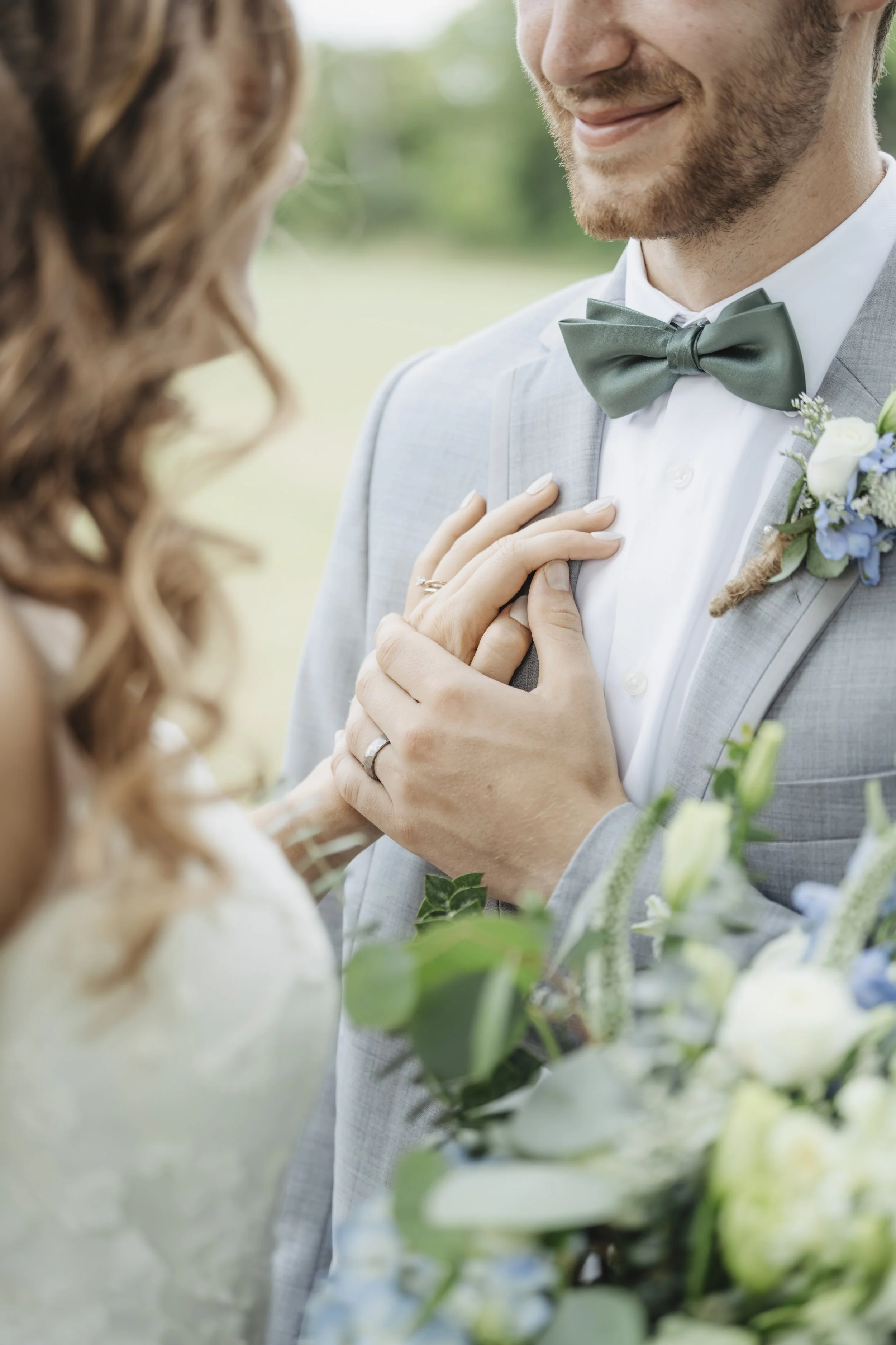 A close-up of a bride and groom at a wedding ceremony. The groom, dressed in a light gray suit with a green bow tie and boutonniere, gently holds the bride's hand over his chest. The bride, partially visible, has long, wavy hair and a wedding dress. 