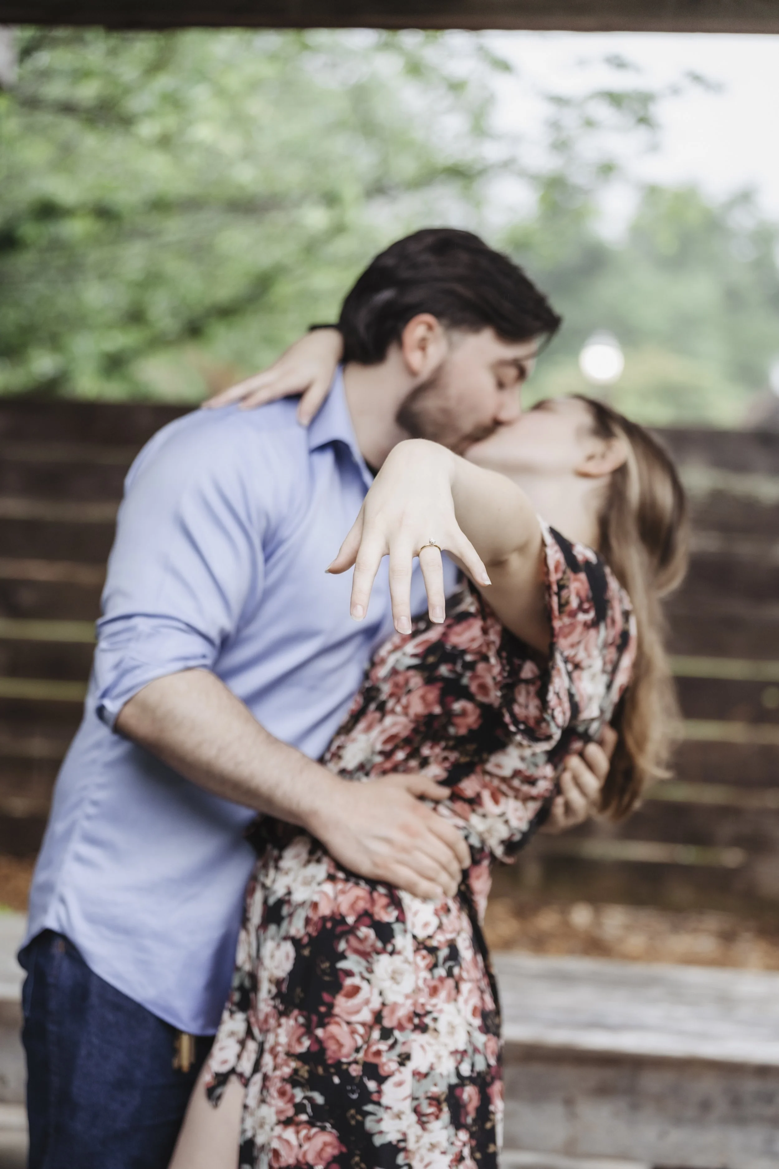 A couple kissing outdoors with the woman displaying a ring on her finger, suggesting an engagement.