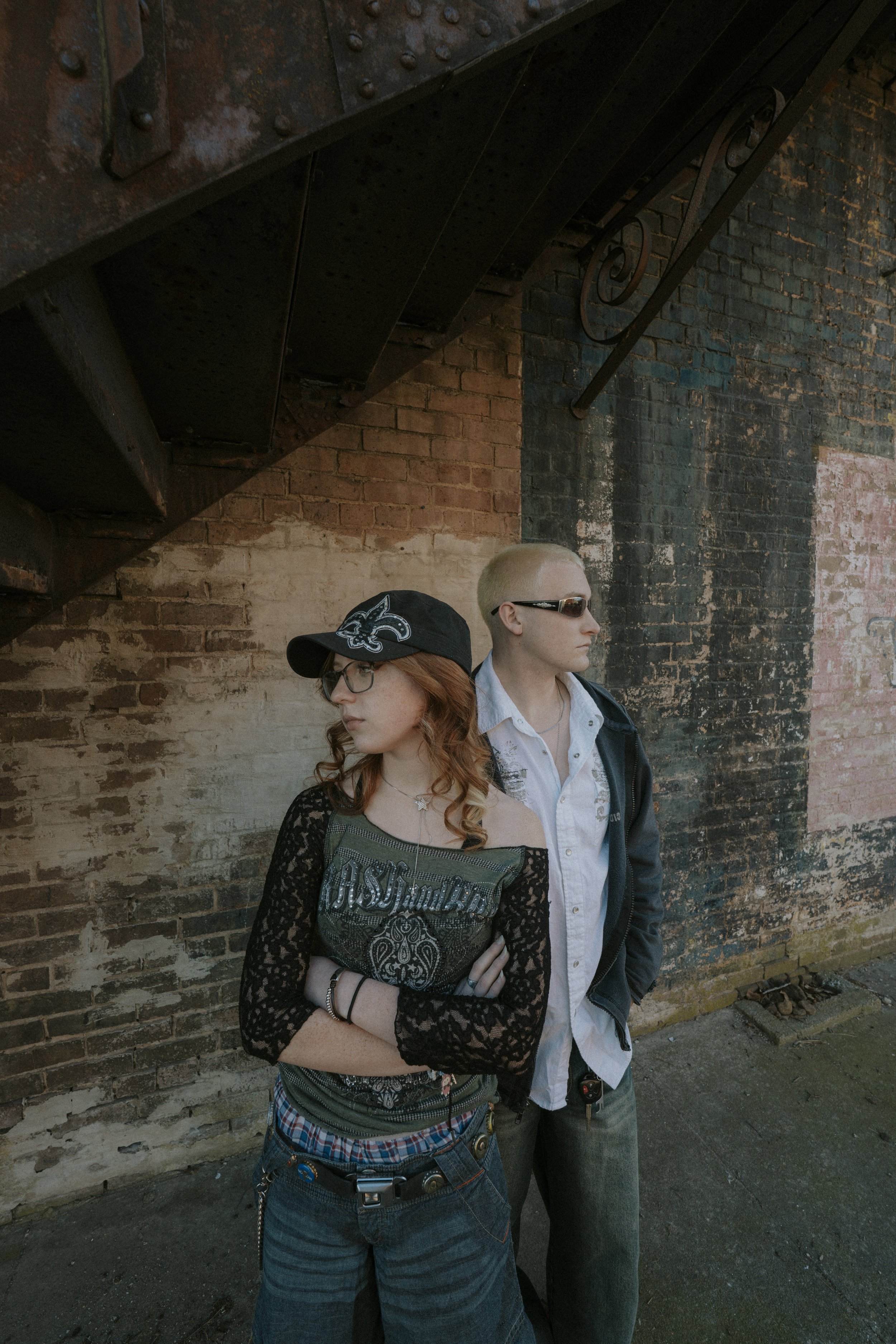 Two young people standing against a weathered brick wall under a brick staircase, both wearing sunglasses and casual clothing, with the woman crossing her arms and the man standing with his hands in his pockets.
