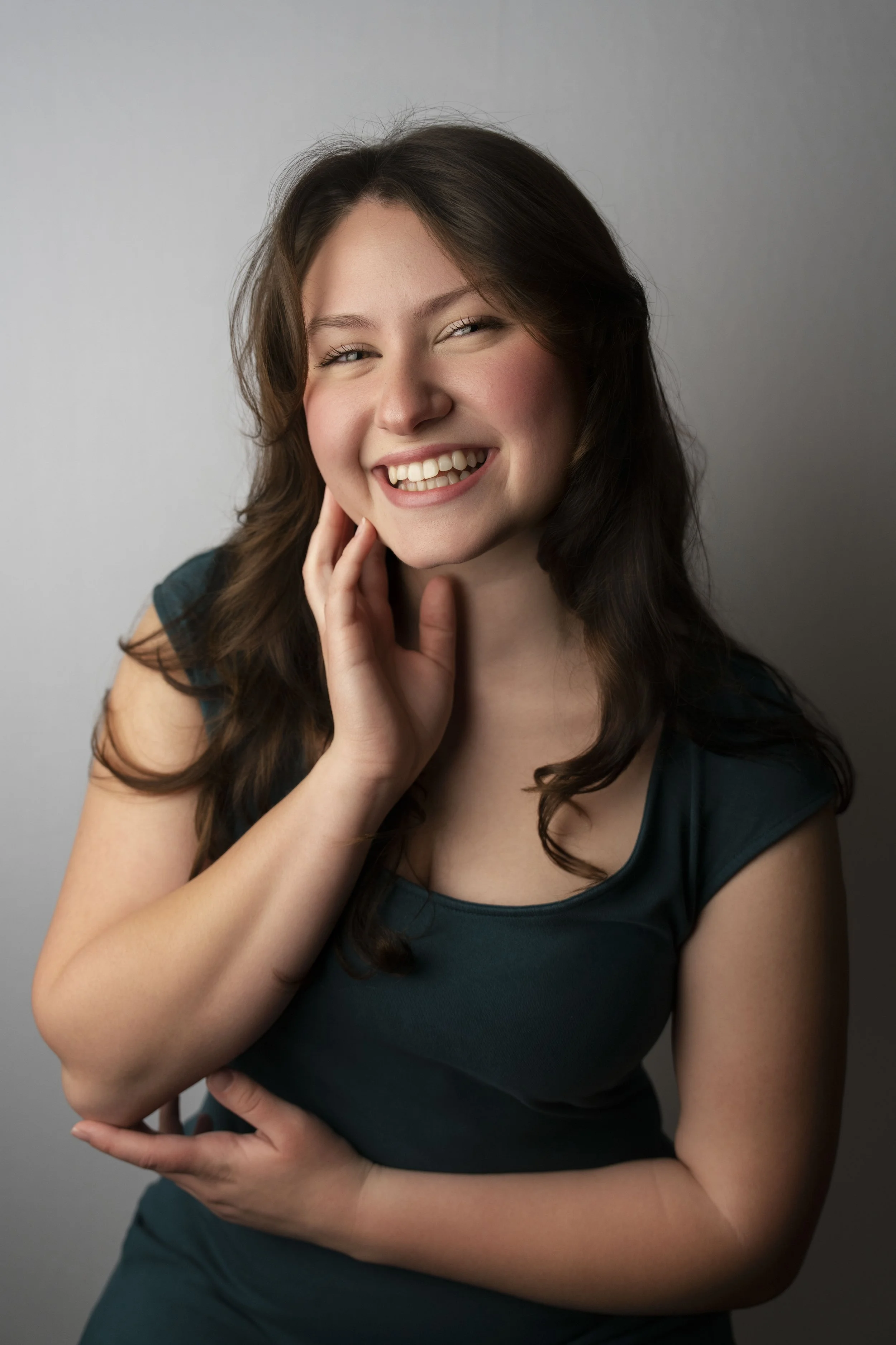 Young woman with long brown hair smiling, touching her face, wearing a teal top, against a plain background.