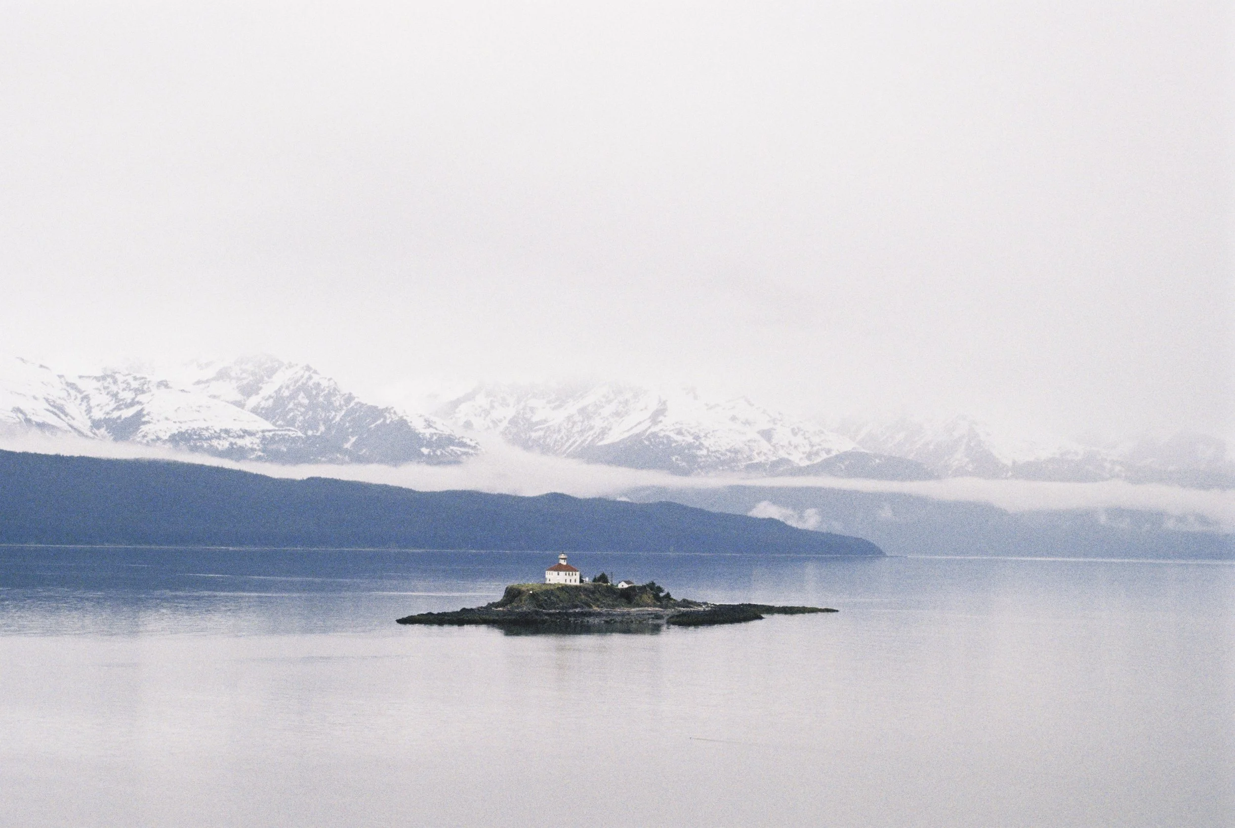 A small lighthouse on a rocky island in calm water, with snow-capped mountains and fog in the background.