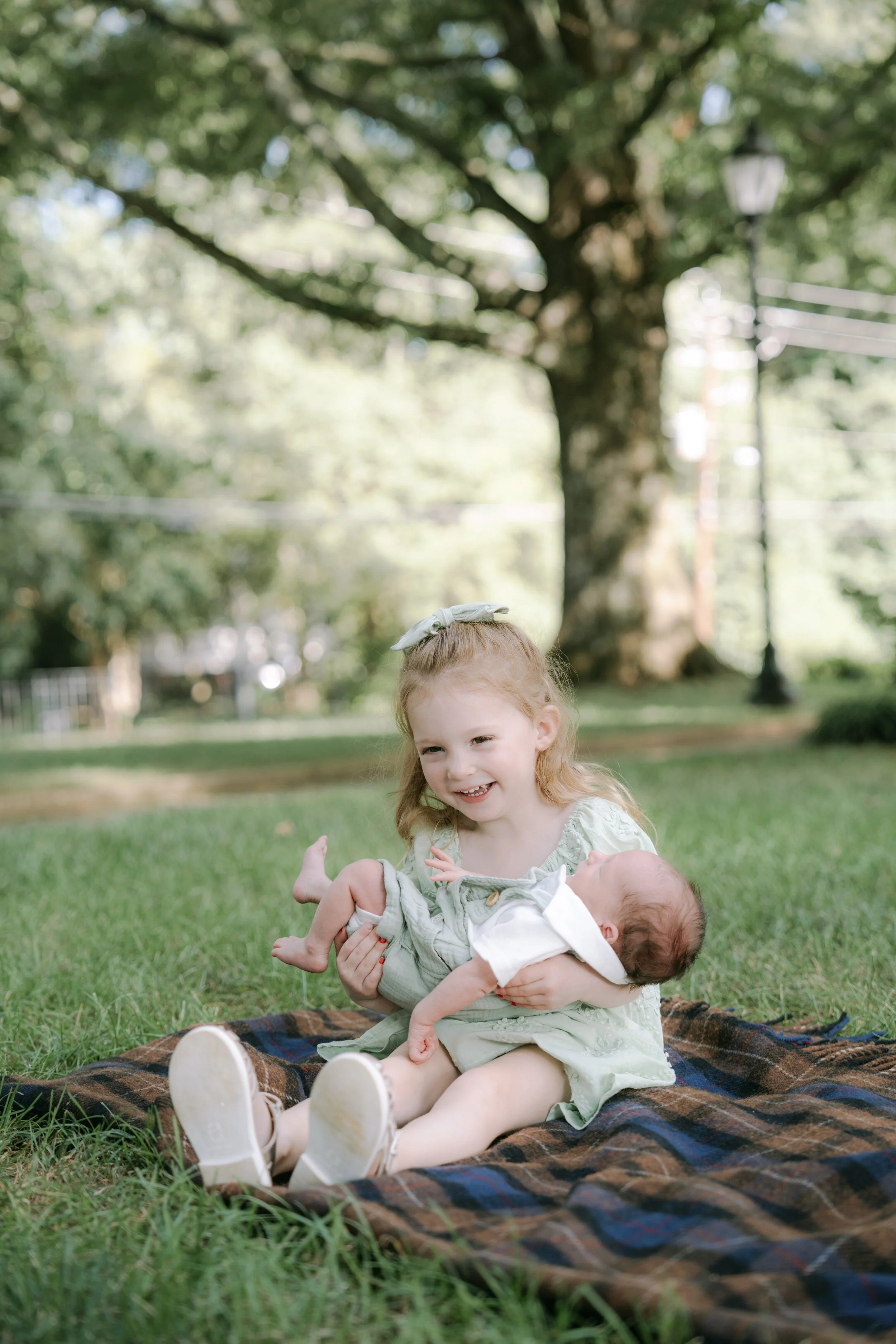 Two young children, a girl and a baby, playing on a blanket outdoors in a park with lush green grass and trees.