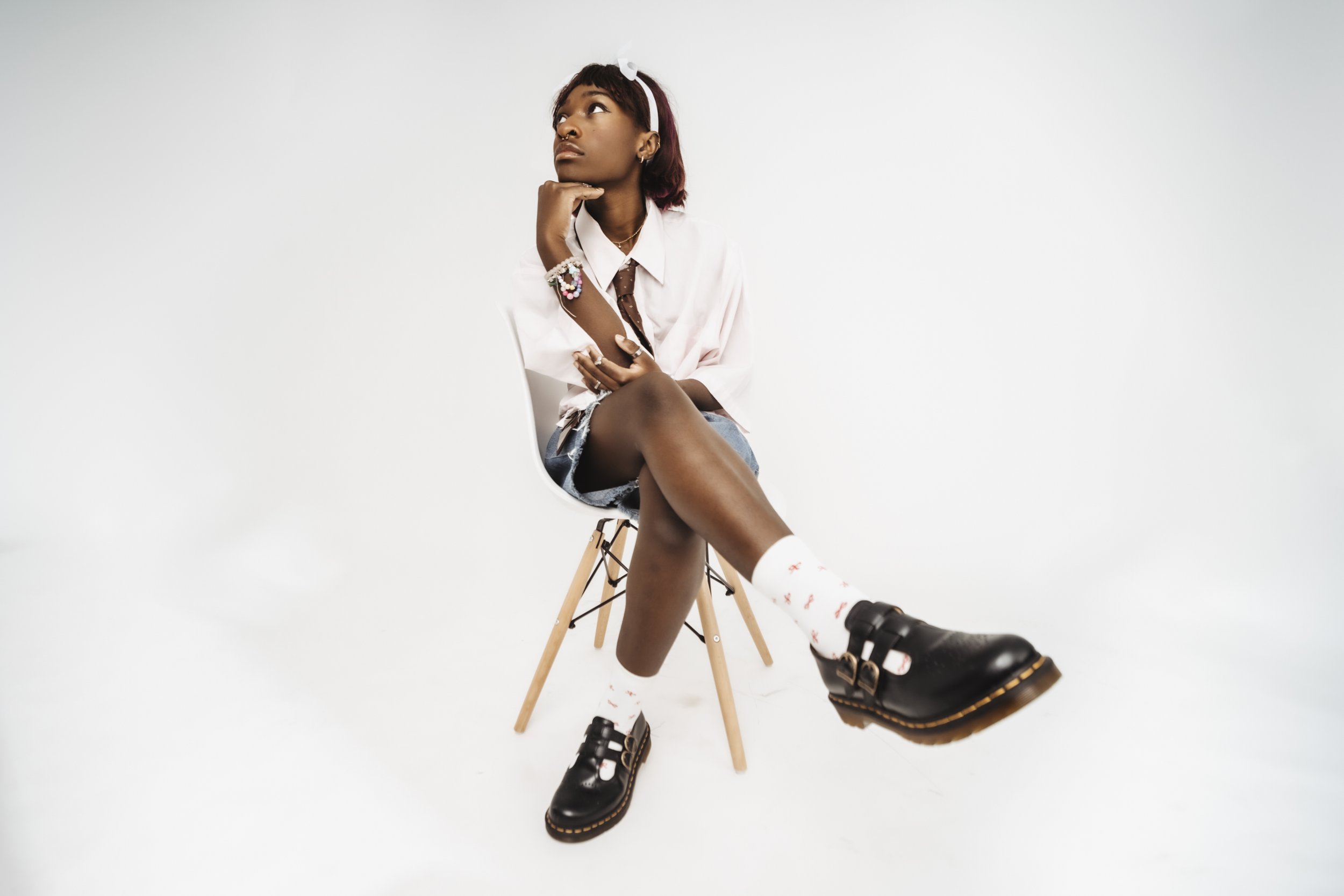 Young woman with dark skin, sitting on a wooden chair against a plain white background, wearing a white shirt, denim shorts, white socks with red patterns, and black shoes, with her hand resting on her chin and looking upwards.