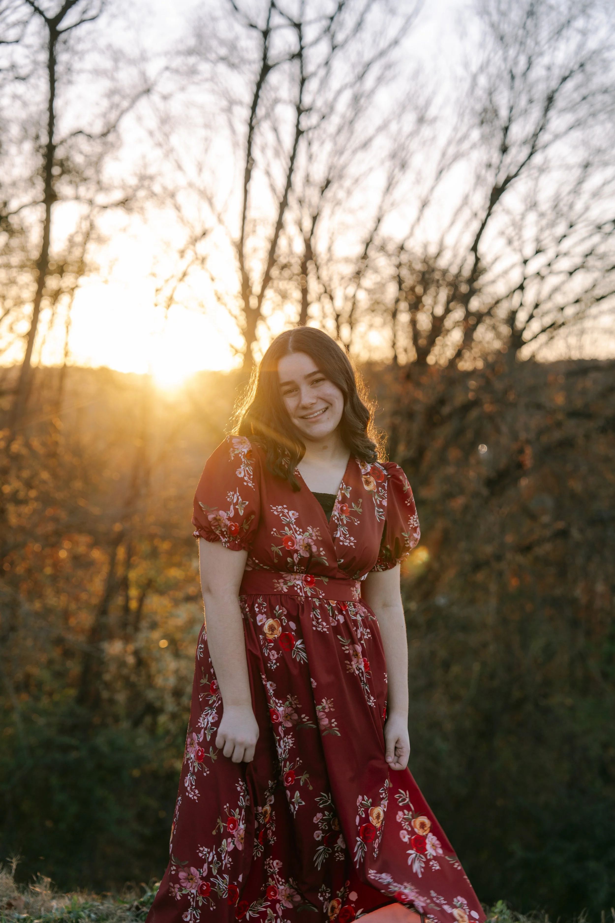 A young woman in a red floral dress standing outdoors during sunset with trees in the background.