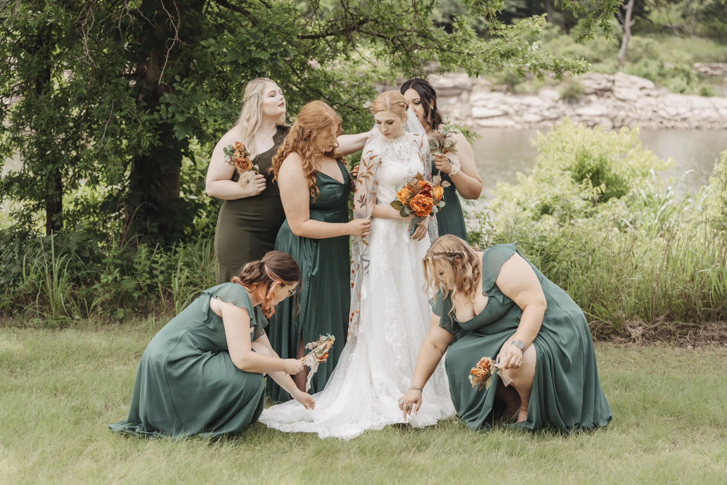 Bridal party outdoors near a river with trees, including a bride in a white gown holding a bouquet of orange flowers, surrounded by bridesmaids in dark green dresses, some bending down adjusting the train of the bride's dress and others standing near