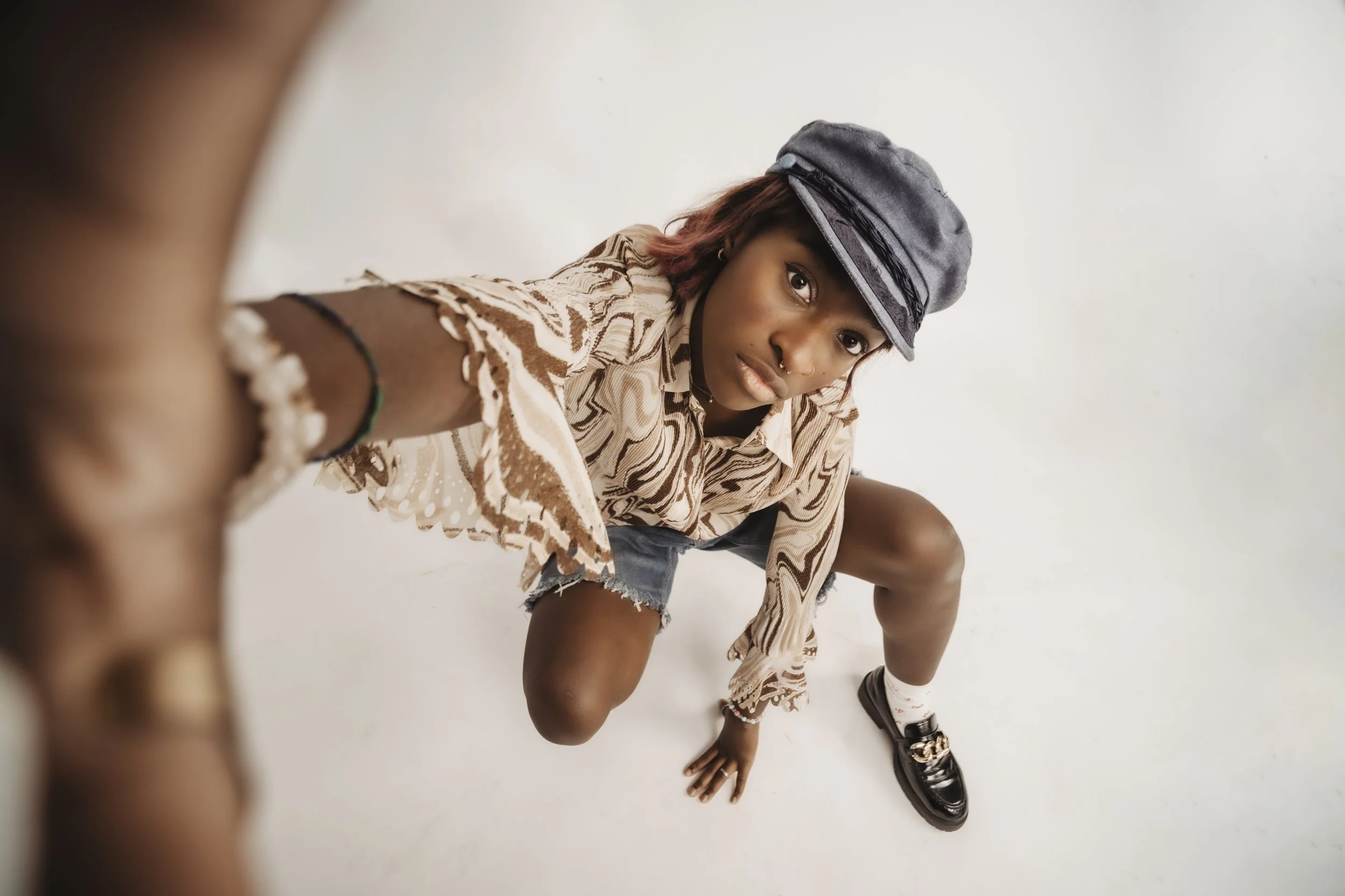 A young woman crouching and looking into the camera, taking a selfie from a low angle, wearing a patterned shirt, denim shorts, a gray cap, and black shoes, against a plain white background.