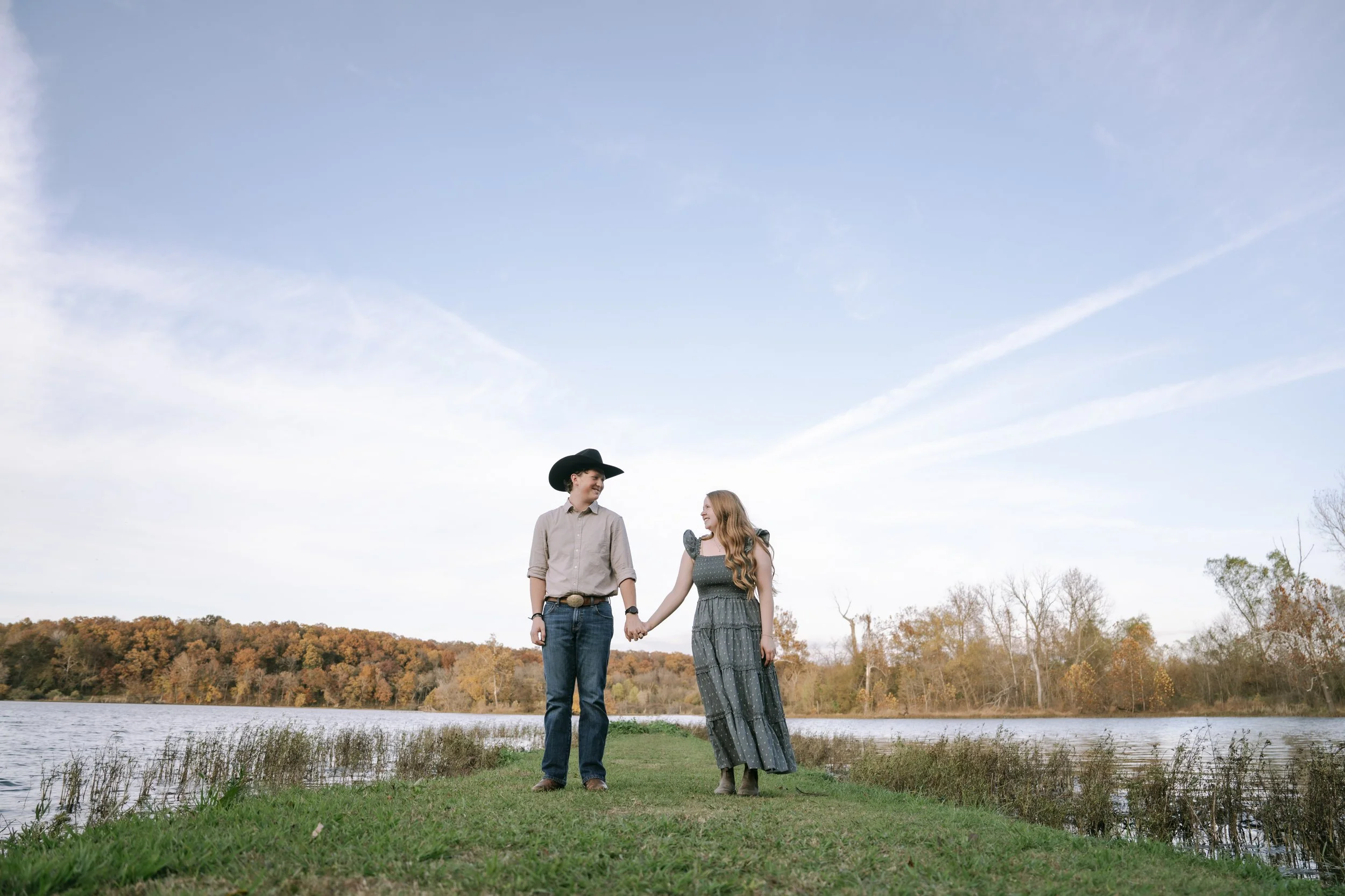 A young couple holding hands on a grassy area near a body of water, with trees showing fall foliage in the background and a blue sky above.