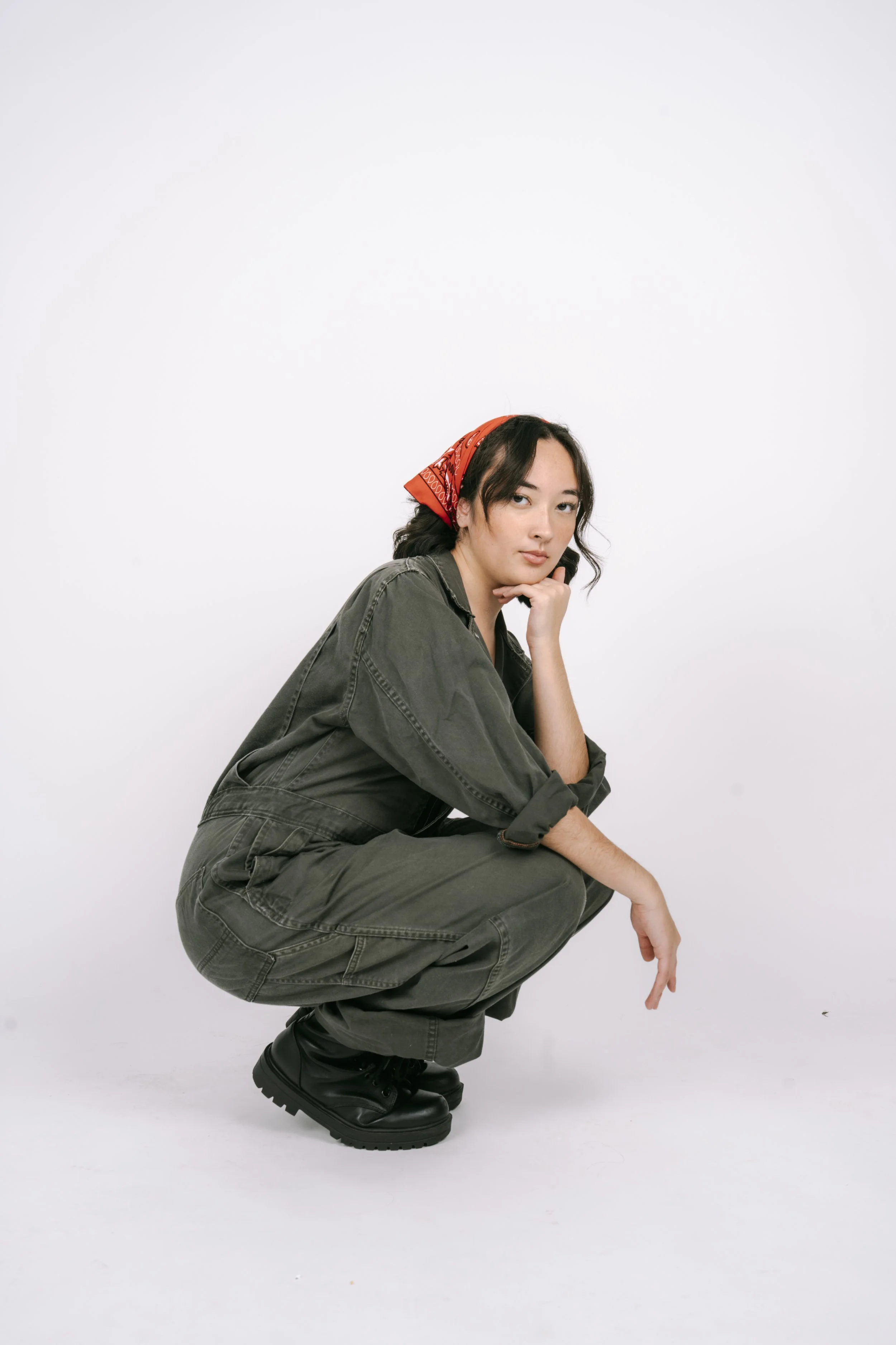 Young woman in dark green jumpsuit and black boots squatting against a plain white background, wearing a red bandana in her hair.